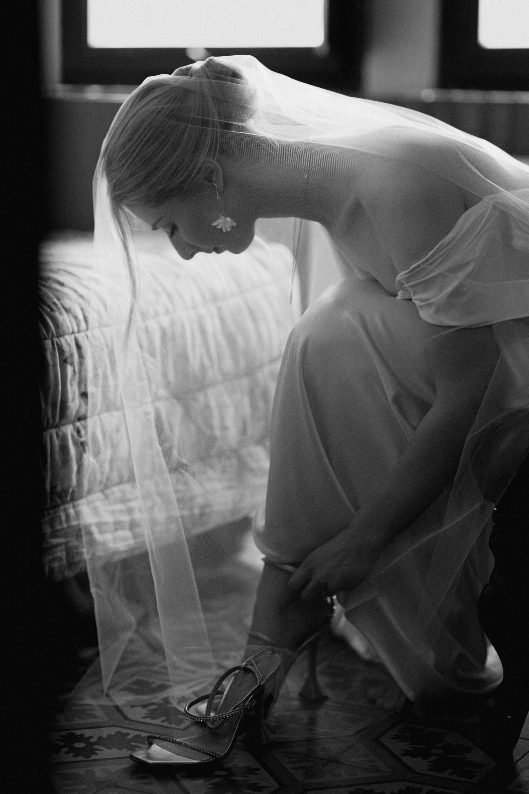 Elegant bride with her hair up in a simple French twist kneeling over her knees to put on her shoes. Photography by Zonzo.
