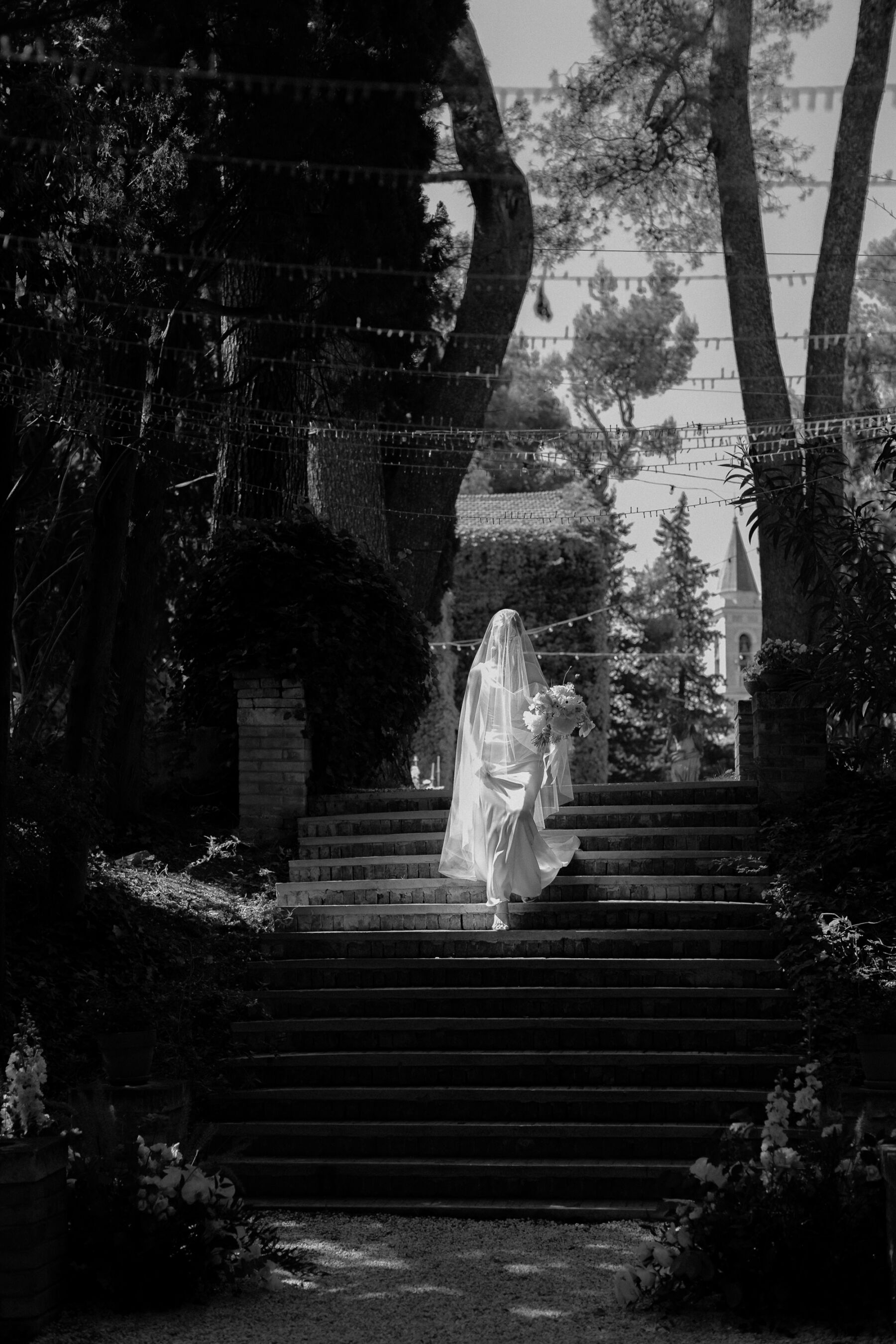 A romantic black and white images of a bride with a veil covering her head, wearing a silk gown by Katherine Tash, descending stone stairs ourdoors on the way to her wedding ceremony. Above her are a canopy of lights that are strung in between the trees that flank the bride's path. Zonzo Photography.
