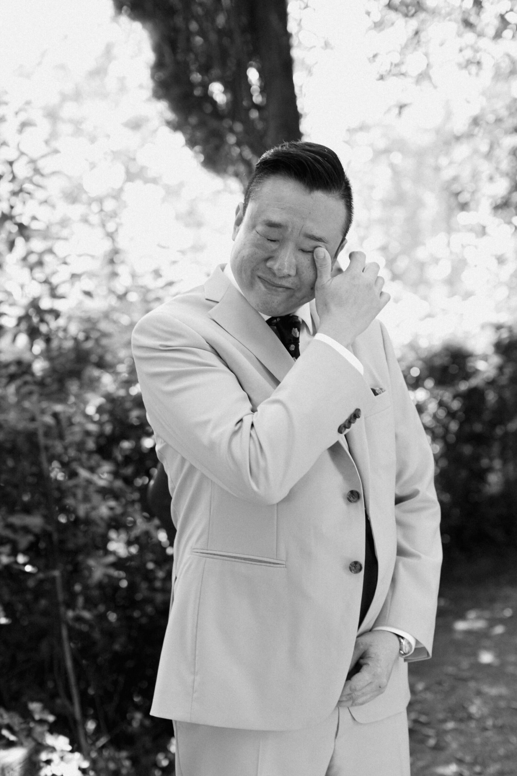 Groom wiping a tear from his eye as he awaits his bride at an outdoor ceremony. Groom wears a pale suit. Zonzo Photography.