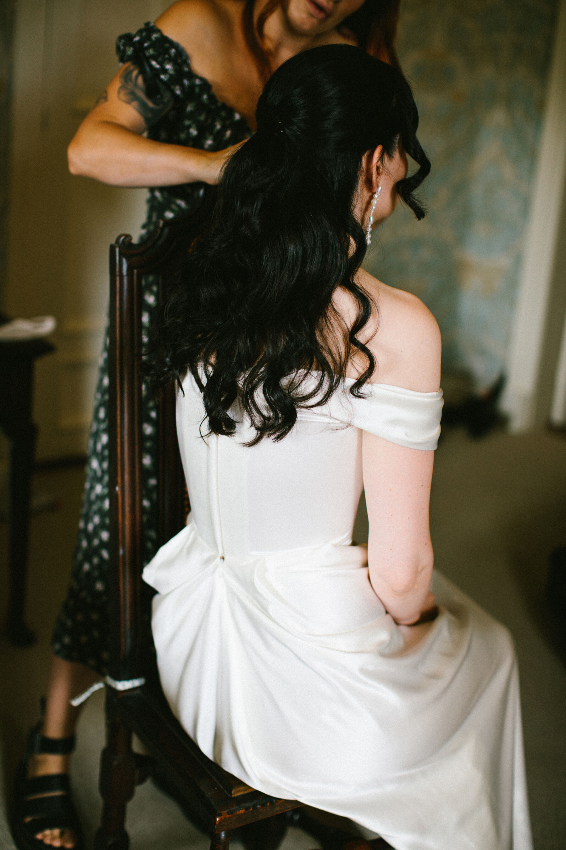 Back shot of a bride sat down having her hair done. She wears a Vivienne Westwood wedding dress, has long dark hair and long pearl earrings.