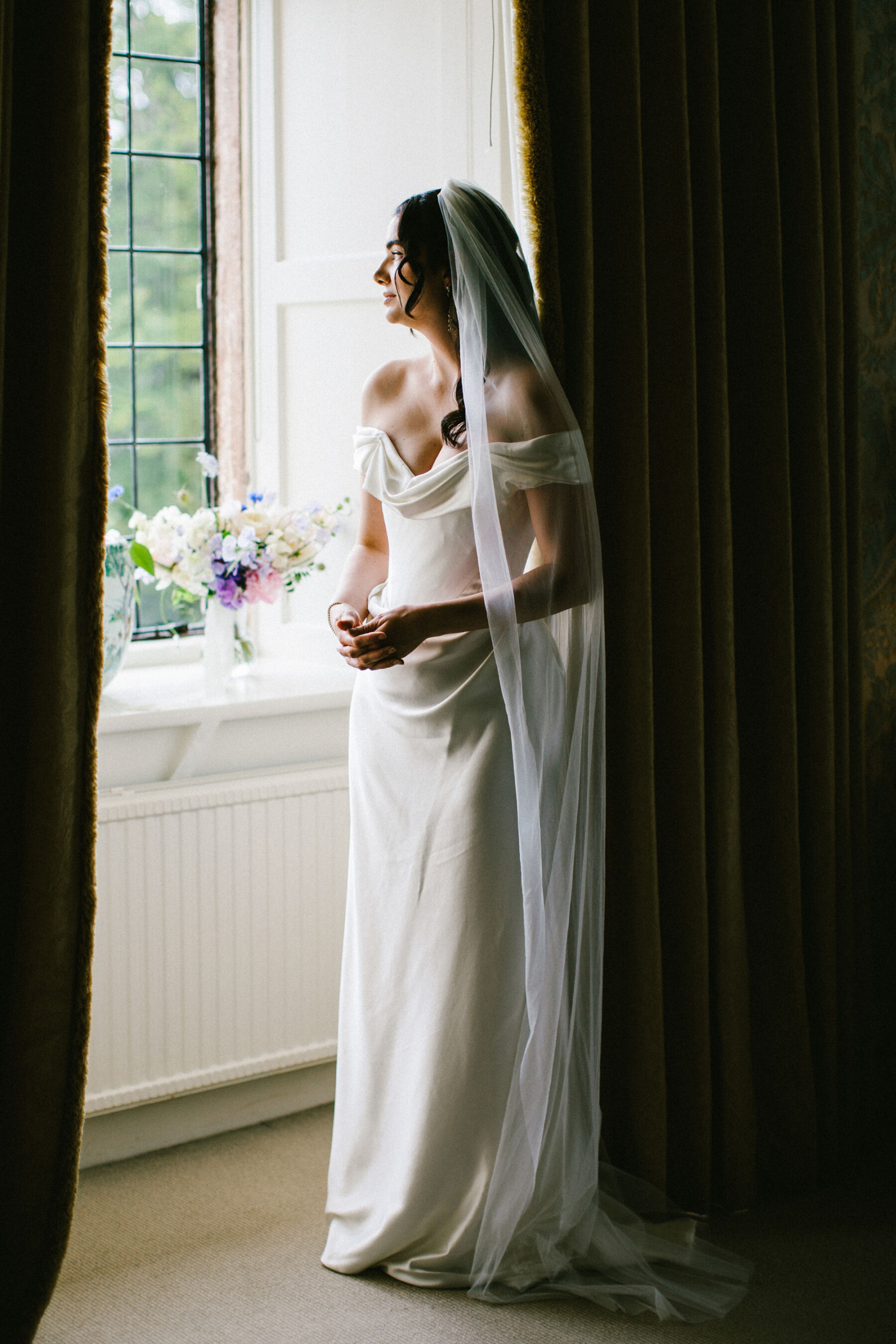 Bride with long dark hair standing next to a window, wearing a Vivienne Westwood wedding dress and a long, single tier wedding veil.