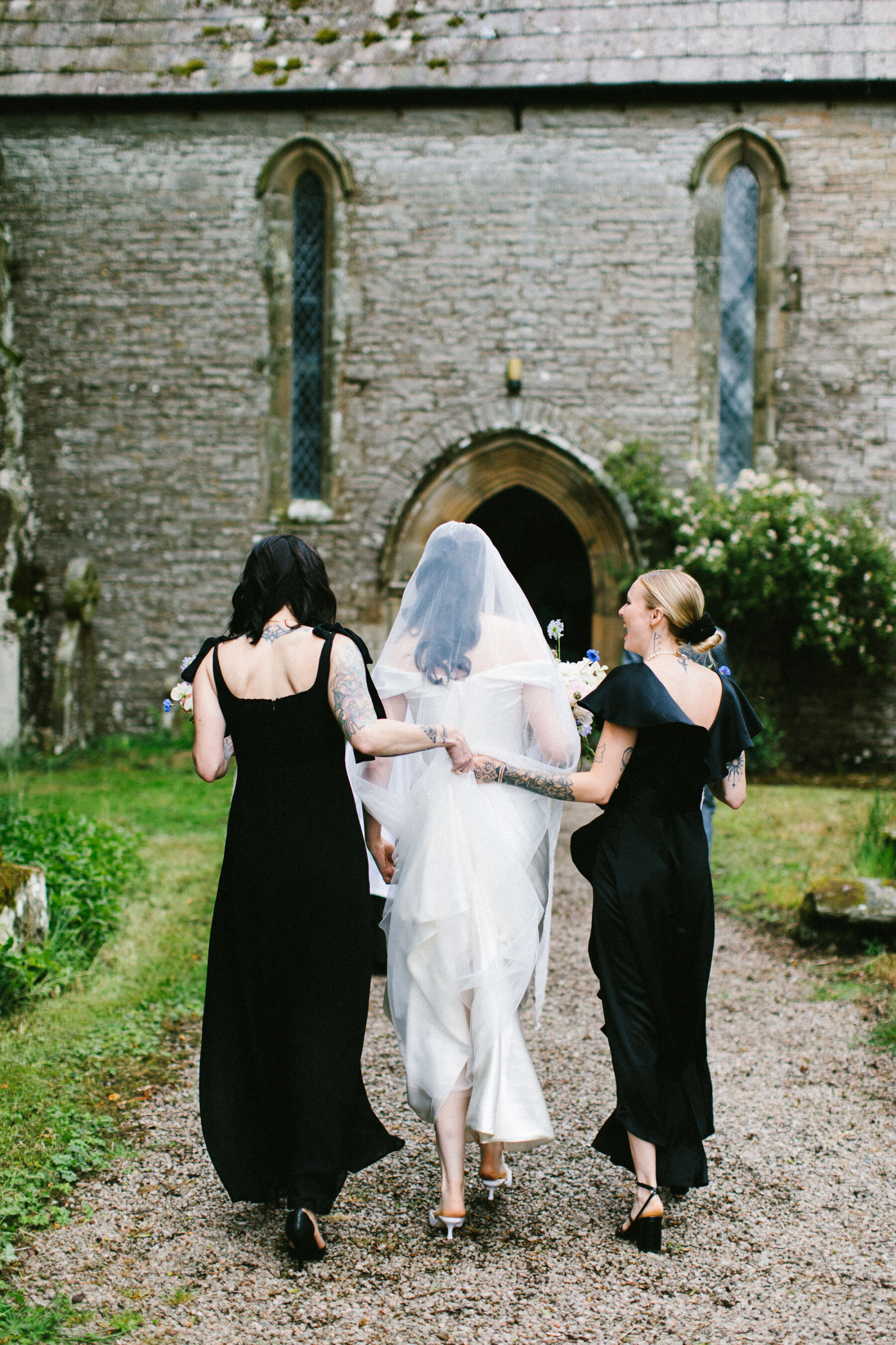 Bridea and her bridesmaids wearing black dresses walking into the church. The bridemaids hold hands behind the bride's back.
