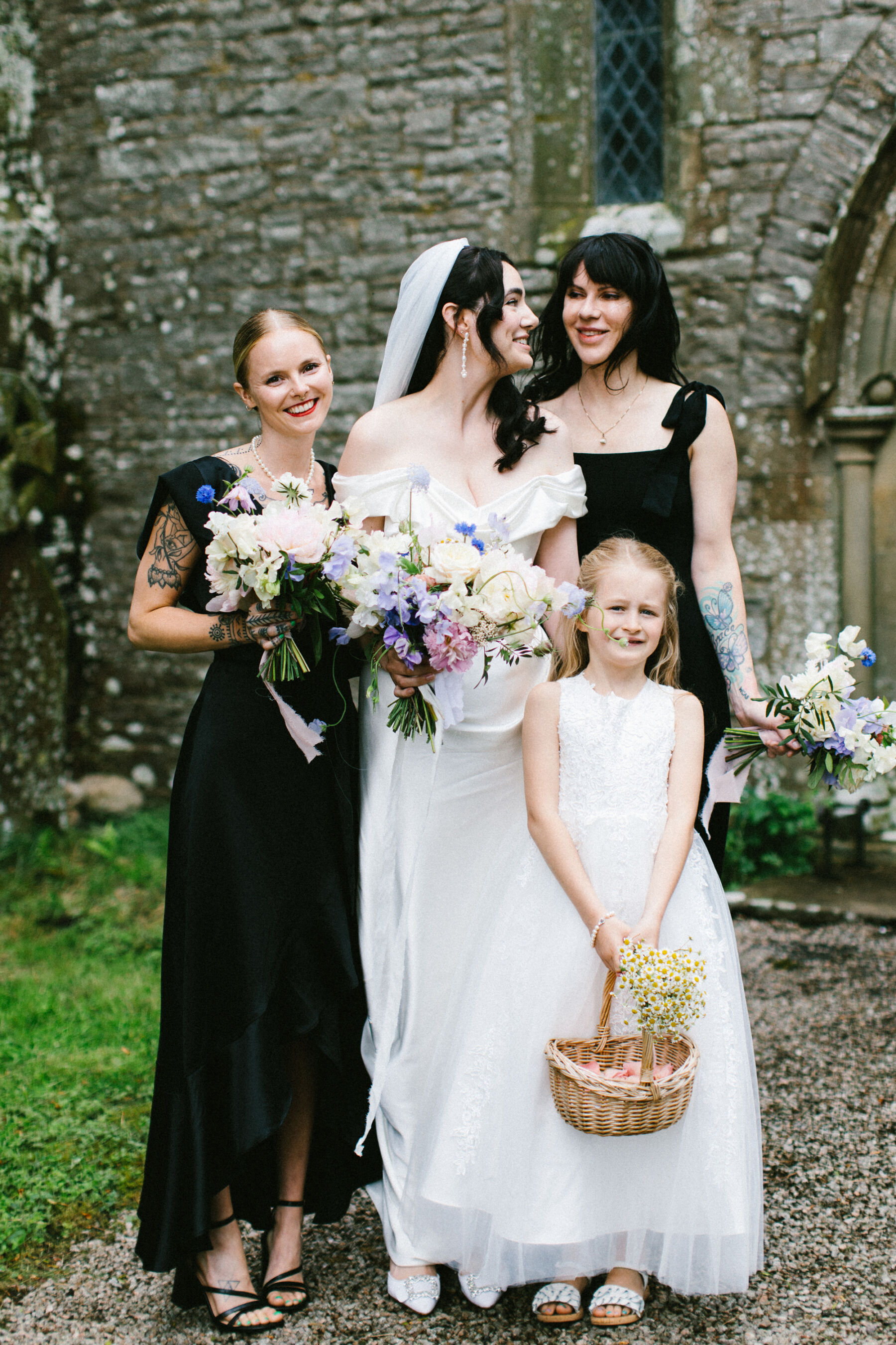 Bride in Vivienne Westwood wedding dress & bridesmaids in black dresses, each carrying elegant summer bouquets. Flowergirl carries a basket of confetti and a bouquet of chamomile.