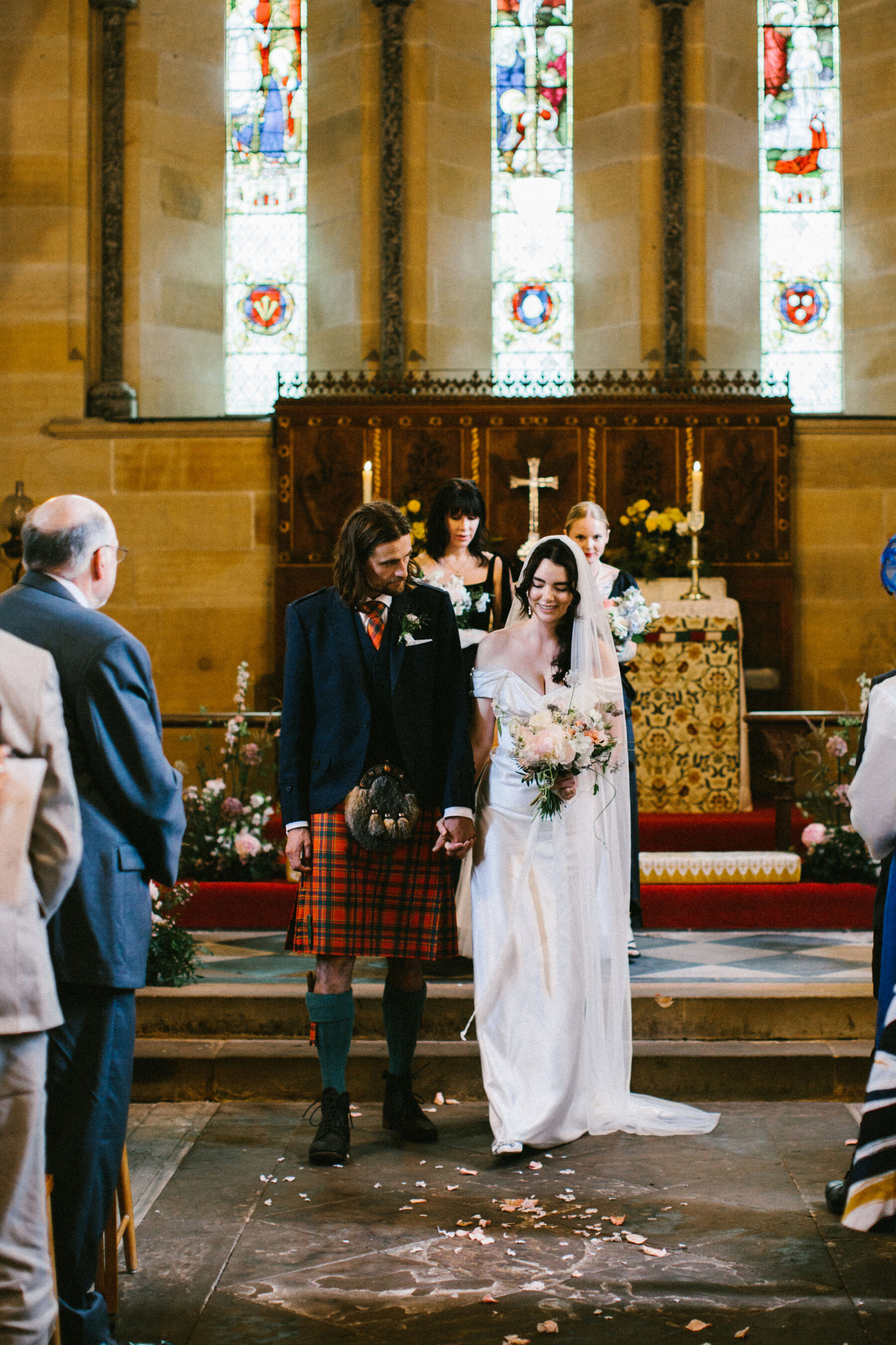 Groom in a red tartan kilt holding hands with his bride in a Vivienne Westwood dress in church.