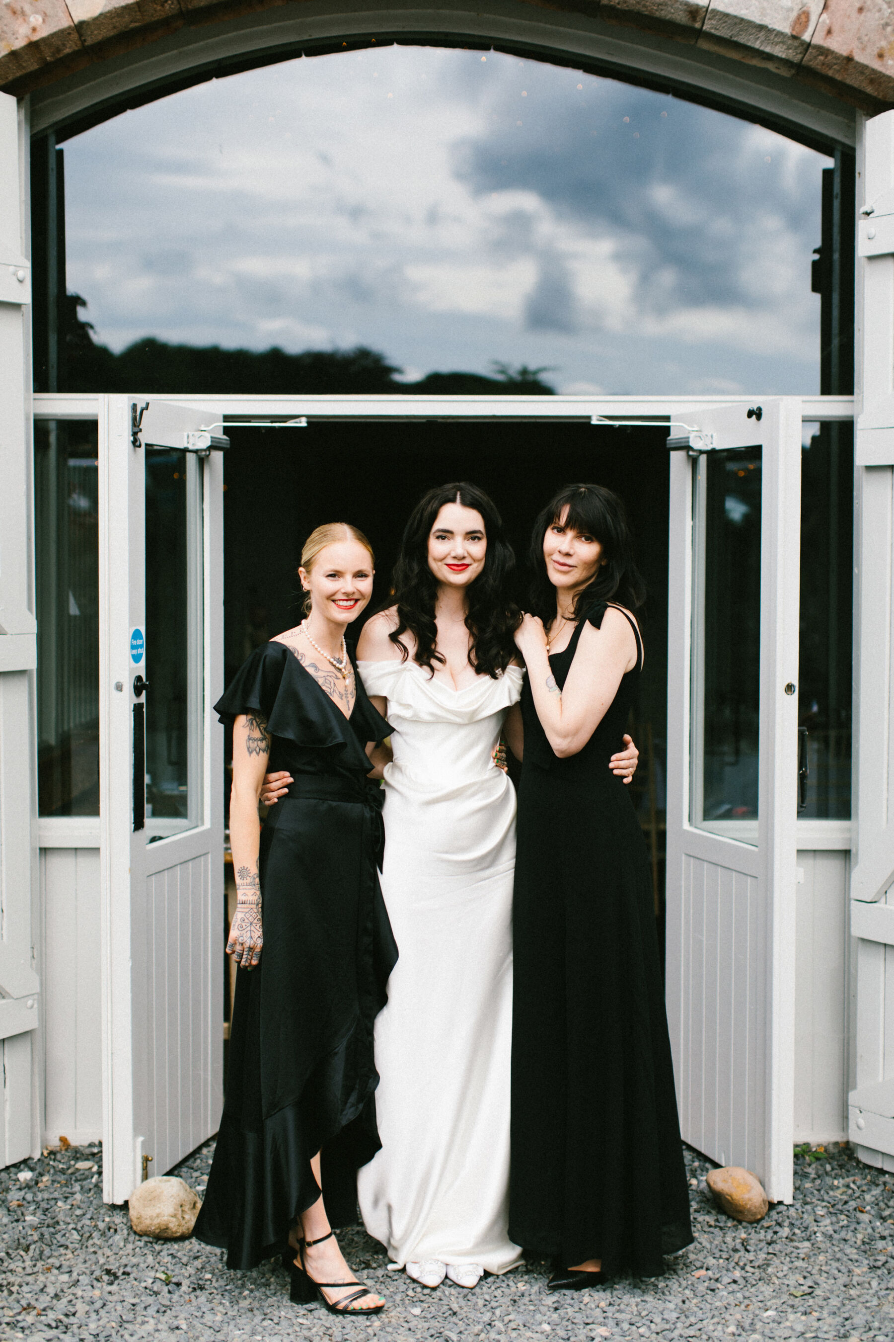 Bride in a Vivienne Westwood wedding dress being flanked by her two bridesmaids in black dresses.