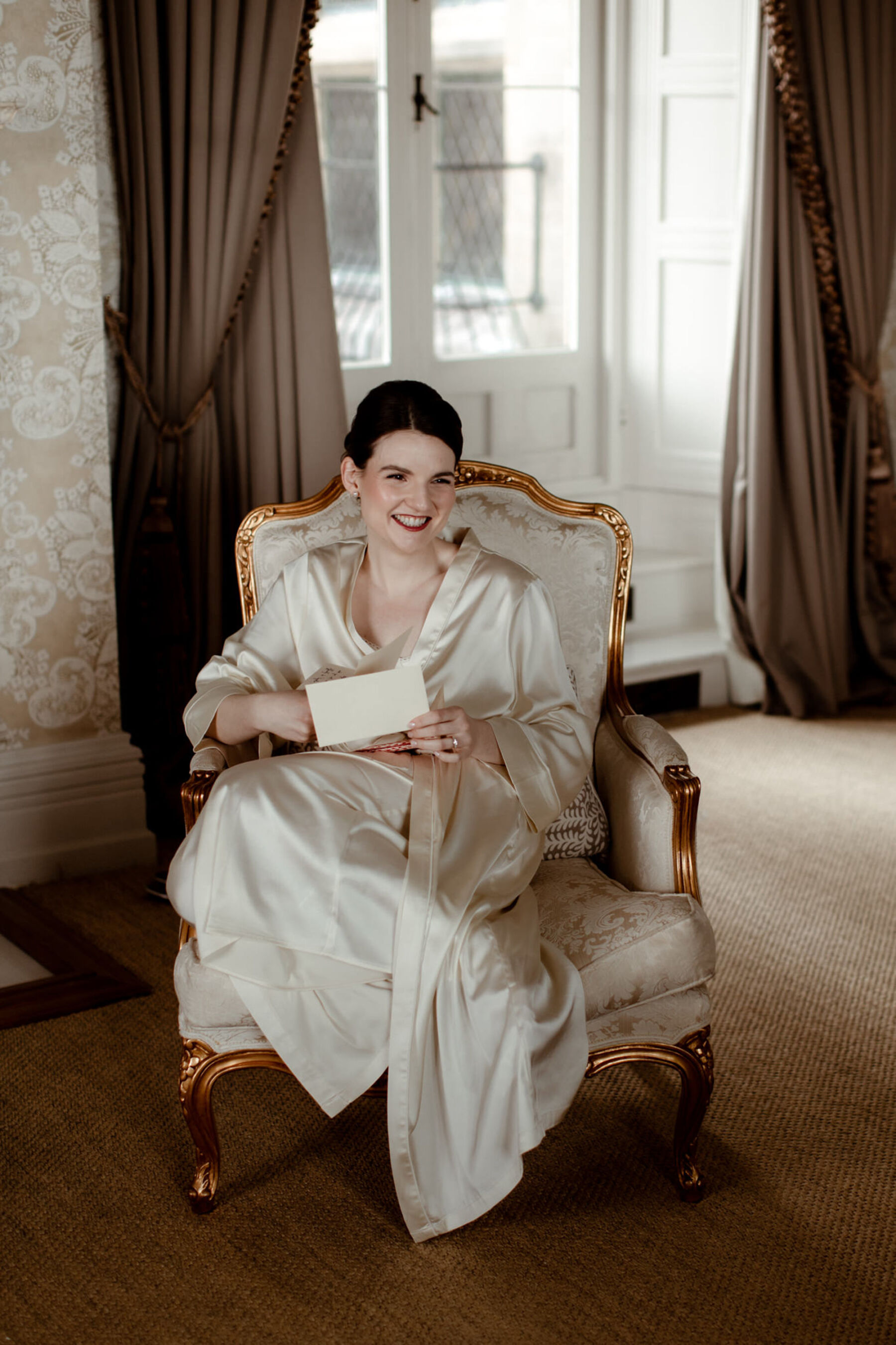Bride in red lipstick and silk pyjamas getting ready on the morning of her wedding. Bride sat smiling on a vintage chair reading a letter from her groom.