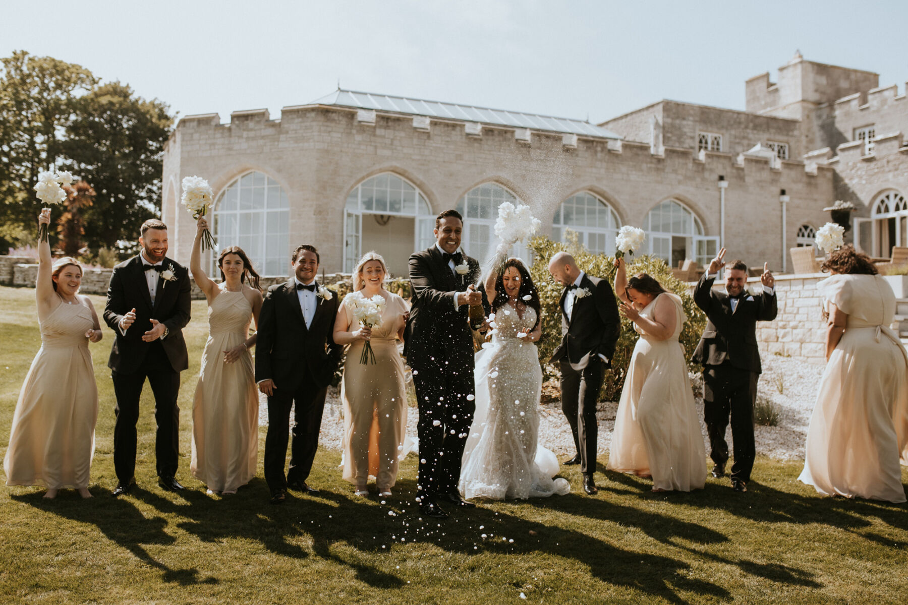 Bride and groom popping a bottle of champagne - surrounded by their bridal party in black tie and pale champagne coloured bridesmaids dresses.