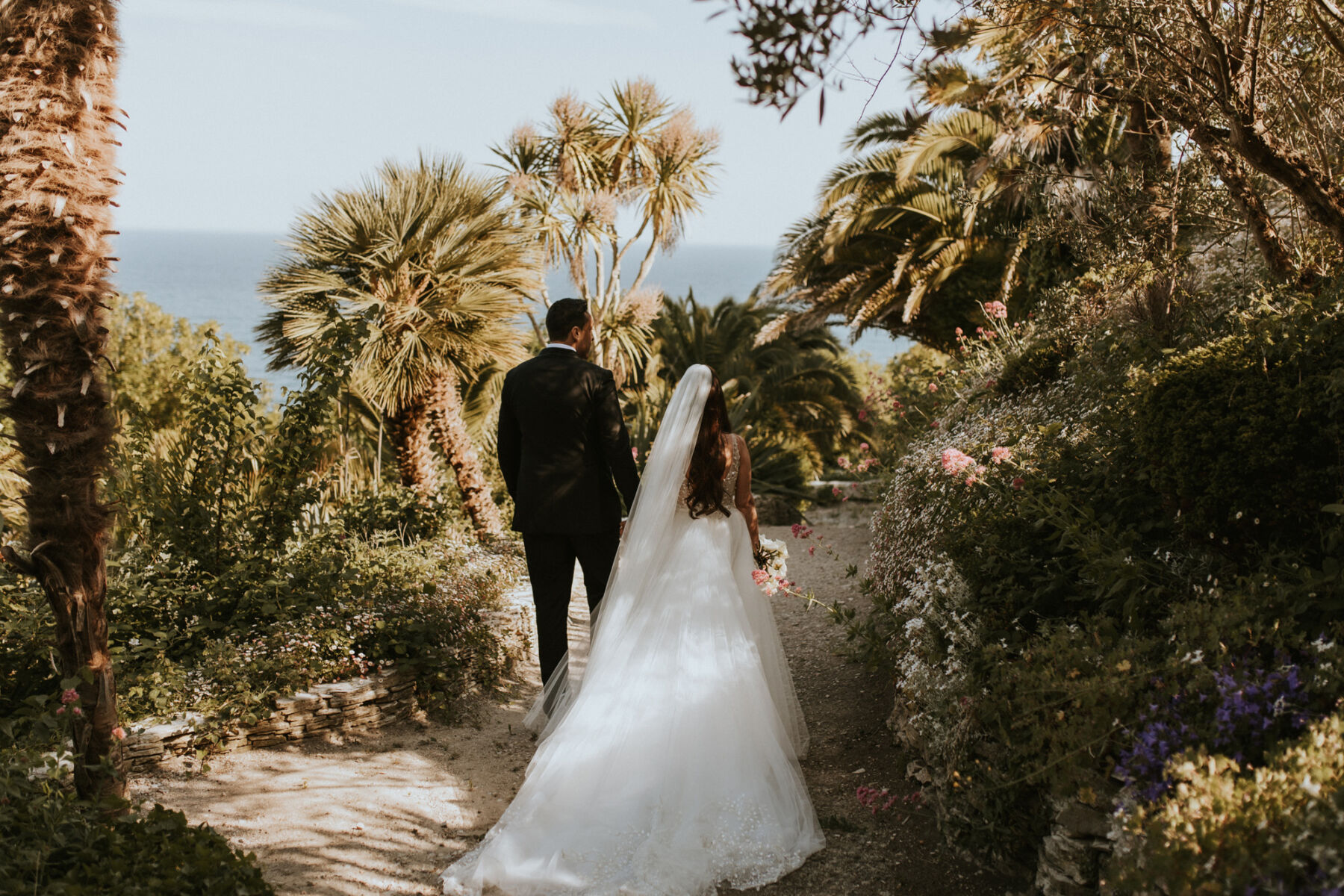 Bride and groom walking down a path surrounded by palm trees at The Pennsylvania Castle Estate - a Dorset wedding venue on a clifftop with incredible sea views.