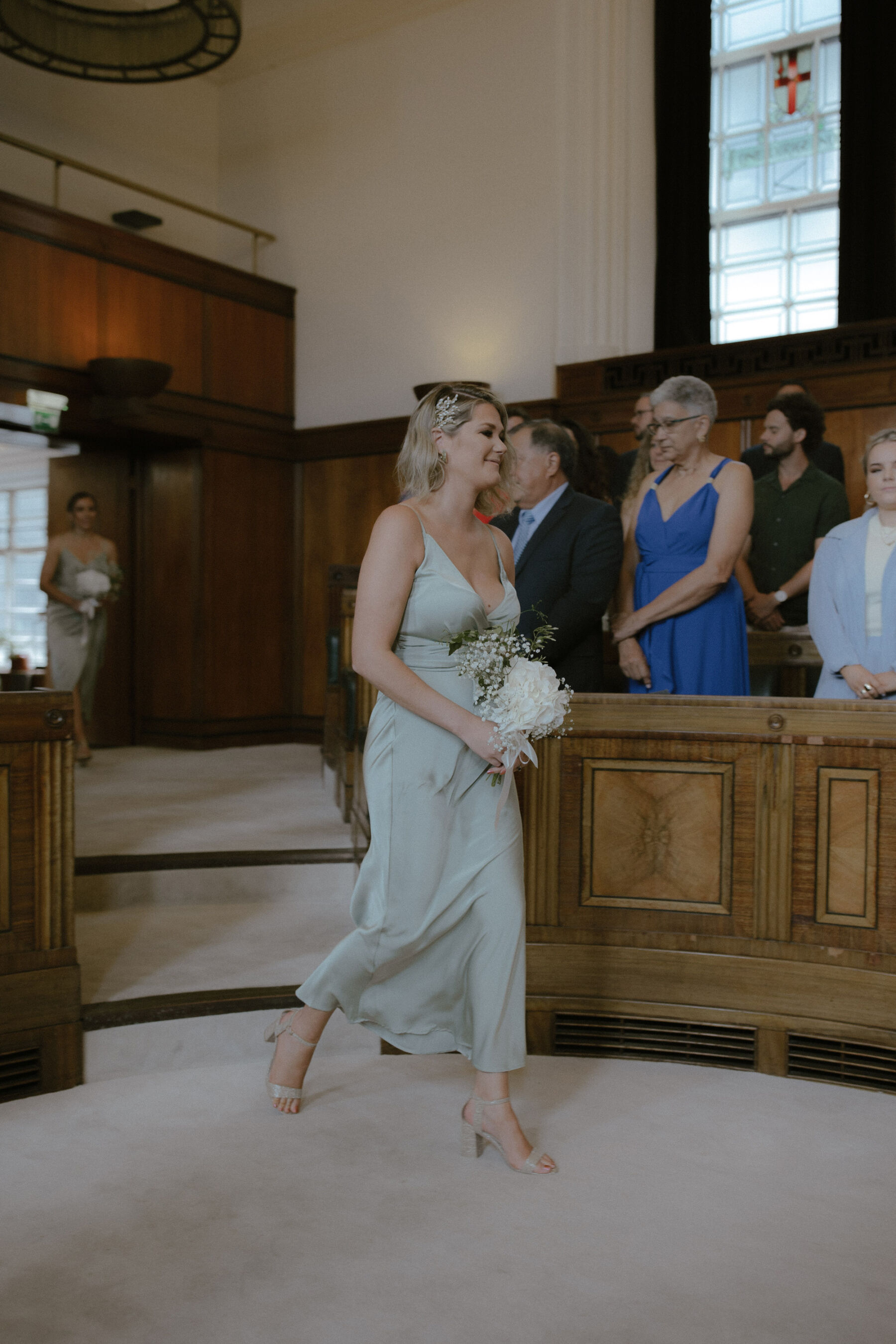 Bride walking into the ceremony wearing a long, sage green dress and carrying a small bouquet of gypsophila and white roses. Ruth Atkinson Photography.