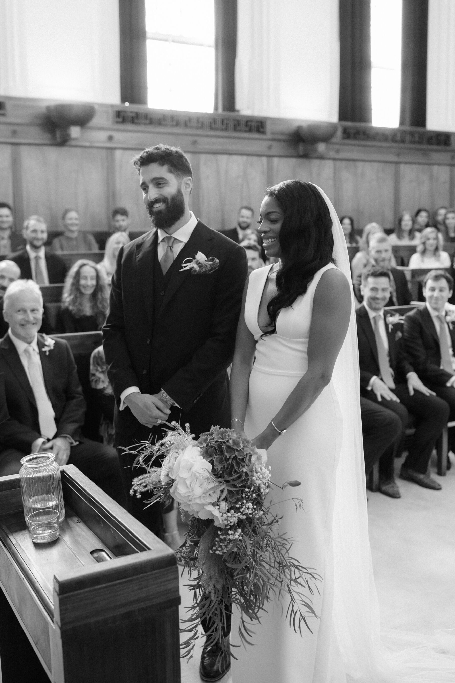 Black and white images of a bride and groom during their wedding ceremony at Town Hall Hotel Bethnal Green. The bride carries a large bouquet and wears a simple, chic & minimalist dress by Sarah Seven. Ruth Atkinson Photography.