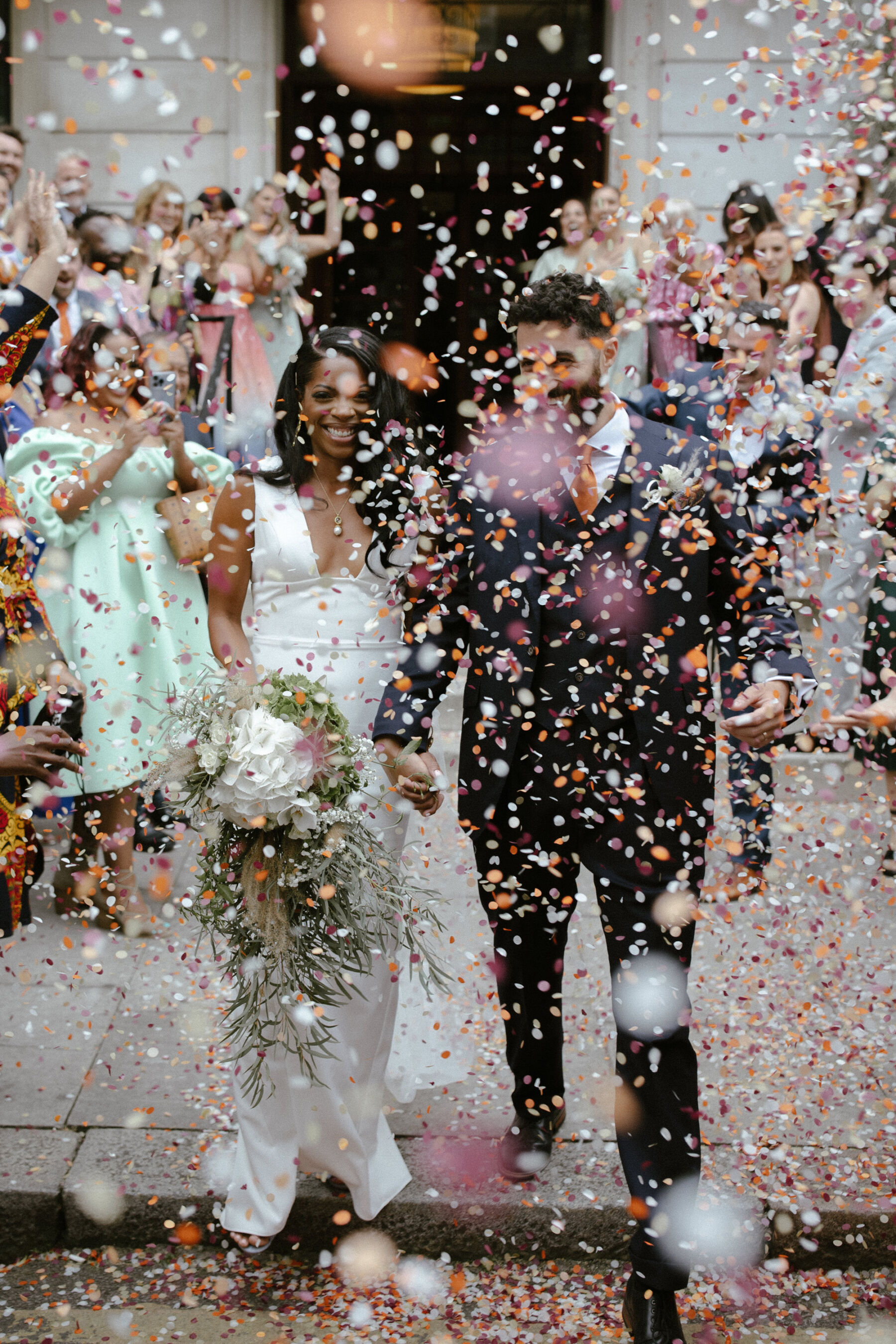 Modern bride and groom shrouded in a cloud of colourful confetti. Bride wears a minimalist dress by Sarah Seven. Ruth Atkinson Photography.