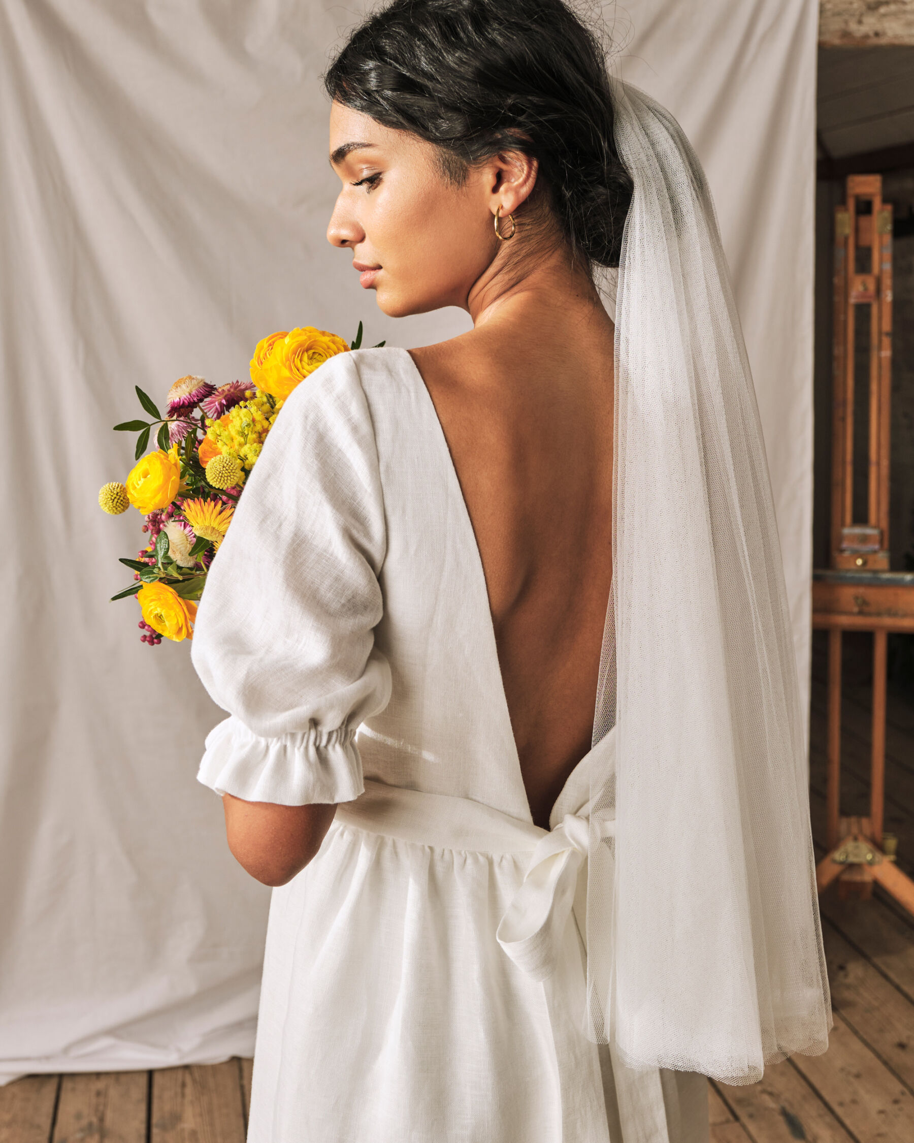 Simple wedding dress with gathered waist and v-back and subtle puff sleeves. Sophie Rose Bridal. Model bride holds a bouquet of yellow flowers.