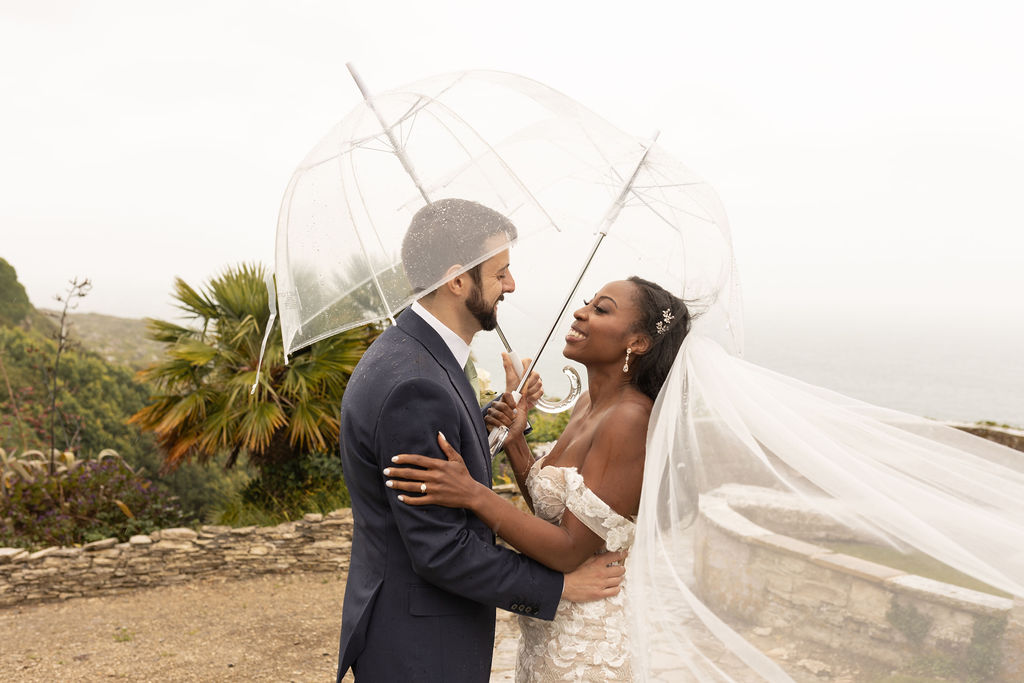 Beautiful Black bride embracing her newlywed husband - both holding umbrellas at The Pennsylvania Castle Estate - a Dorset wedding venue on a clifftop with incredible sea views.