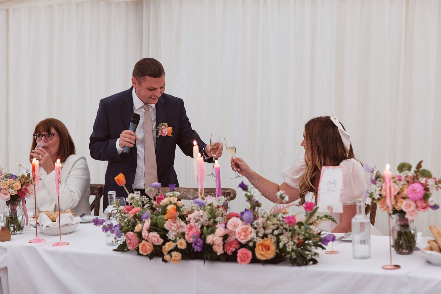 Bride and groom sitting at the top table with pink taper candles, a colourful 'long and low' floral arrangement and chinking their champagne glasses.