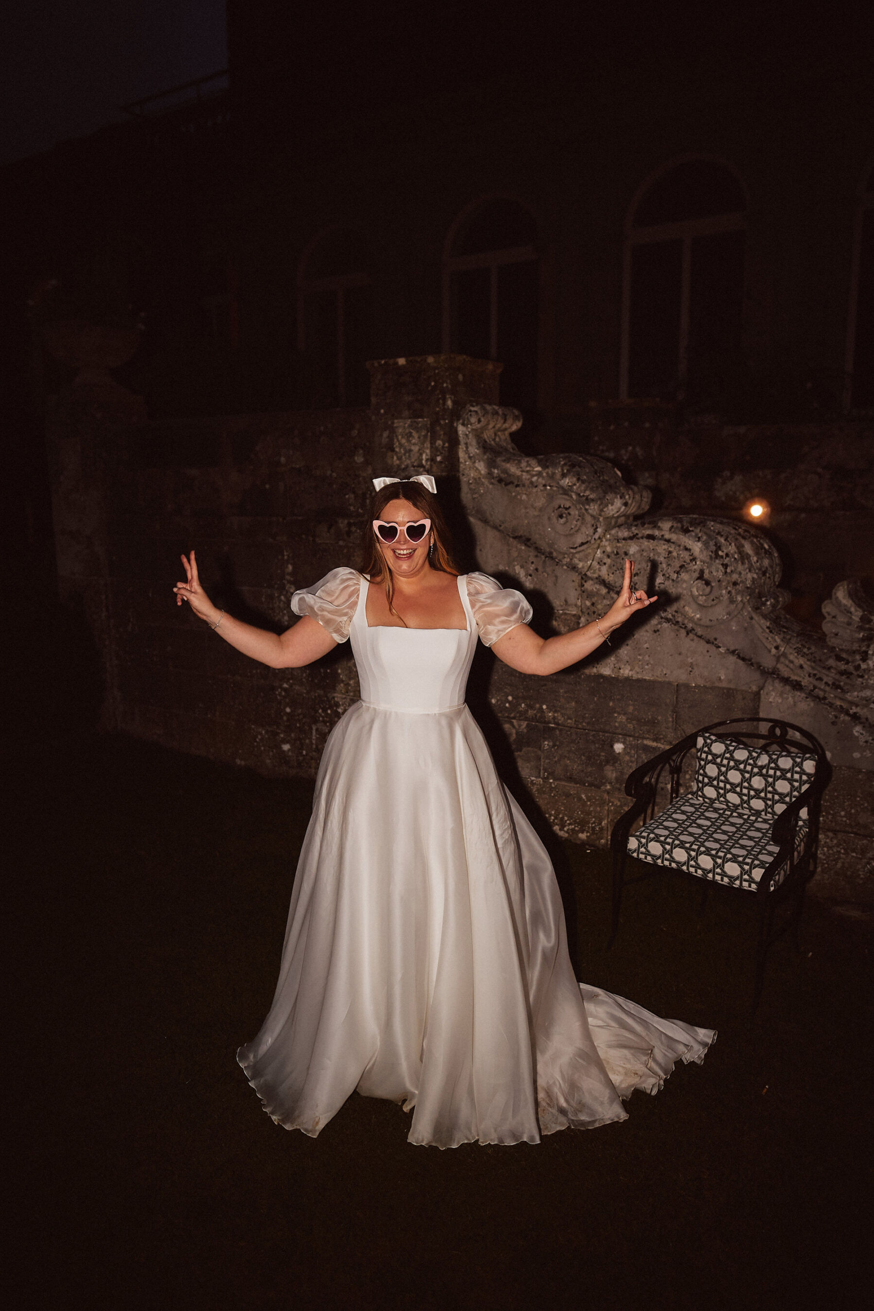 A Romantic, Bridgerton Inspired Wedding at Cowley Manor 33 Bride and groom making peace sign gestures with her hands, wearing heart shaped sun glasses and a puff sleeved Suzanne Neville wedding dress. She wears a Gigi & Olive bow in her hair.