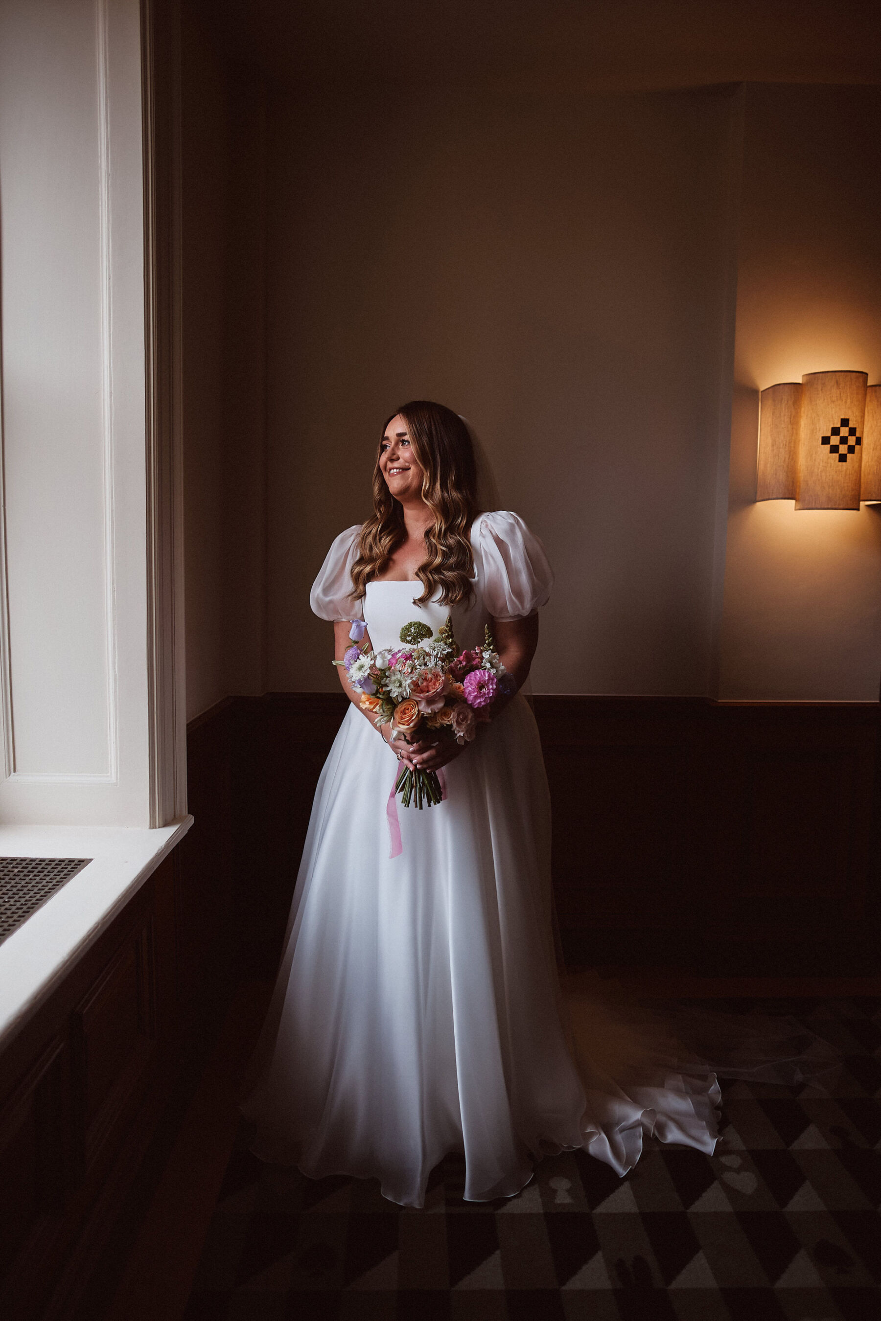 Bride in a puff sleeve Suzanne Neville wedding dress, standing by a window and bathed in window light, and carrying a colourful wedding bouquet.
