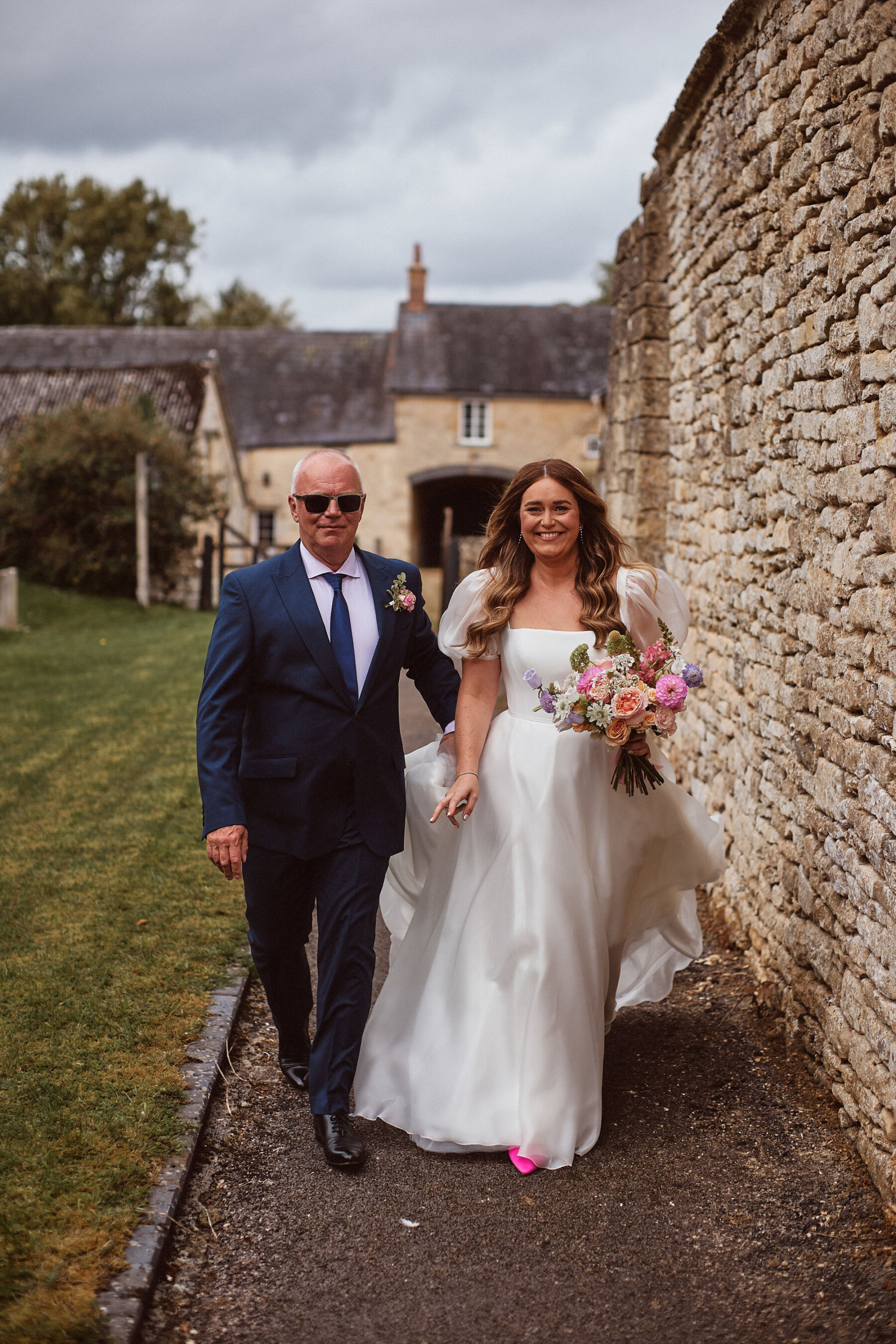 Happy bride with her father, she wears a puff sleeve Suzanne Neville wedding dress and bright pink shoes from New Look. She carries a colourful (pinks & pastels) wedding bouquet.