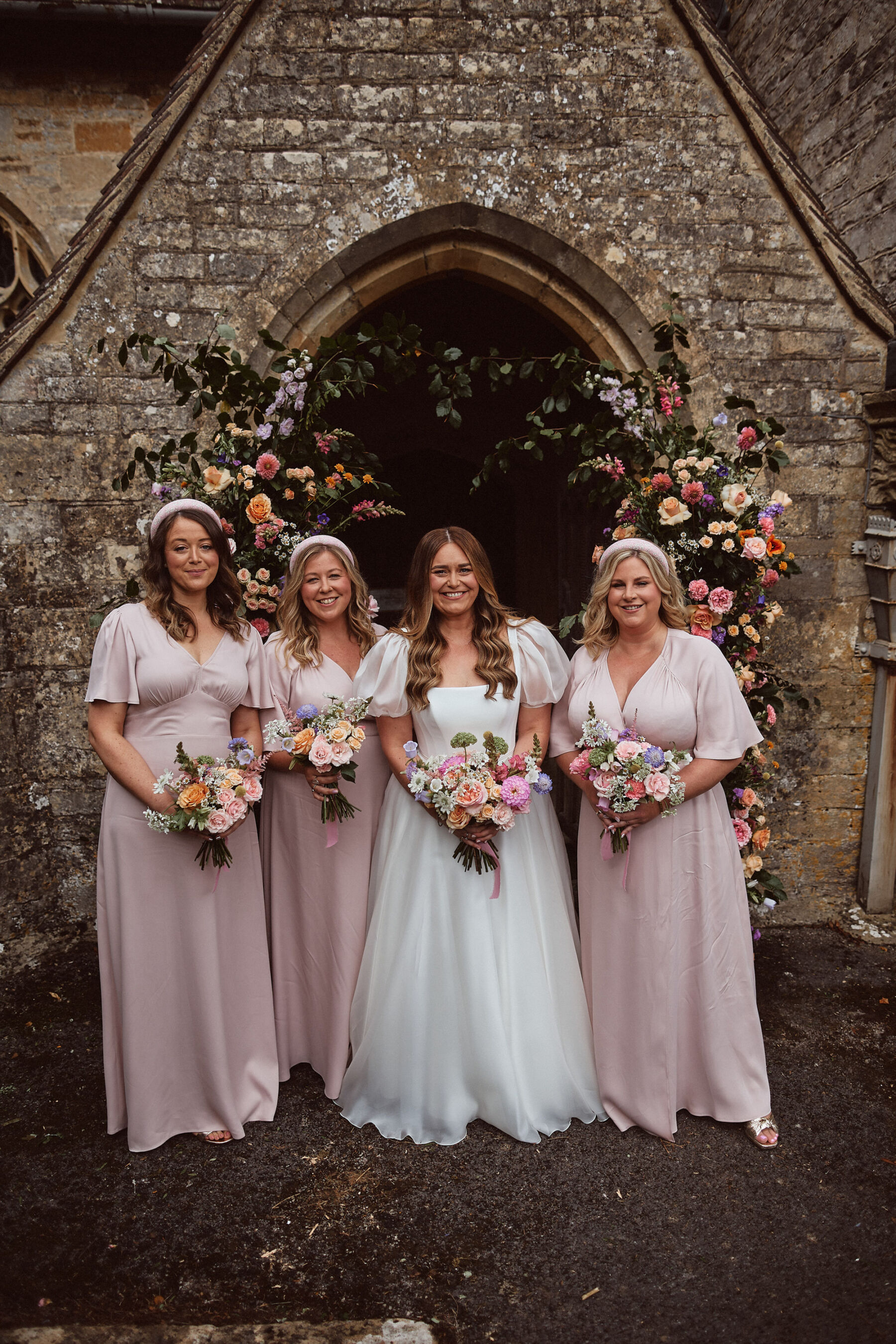 Bridesmaids in pink Maids to Measure bridesmaids dresses, standing with the bride in a puff sleeve Suzanne Neville dress buy a church door decorated with colourful flowers.