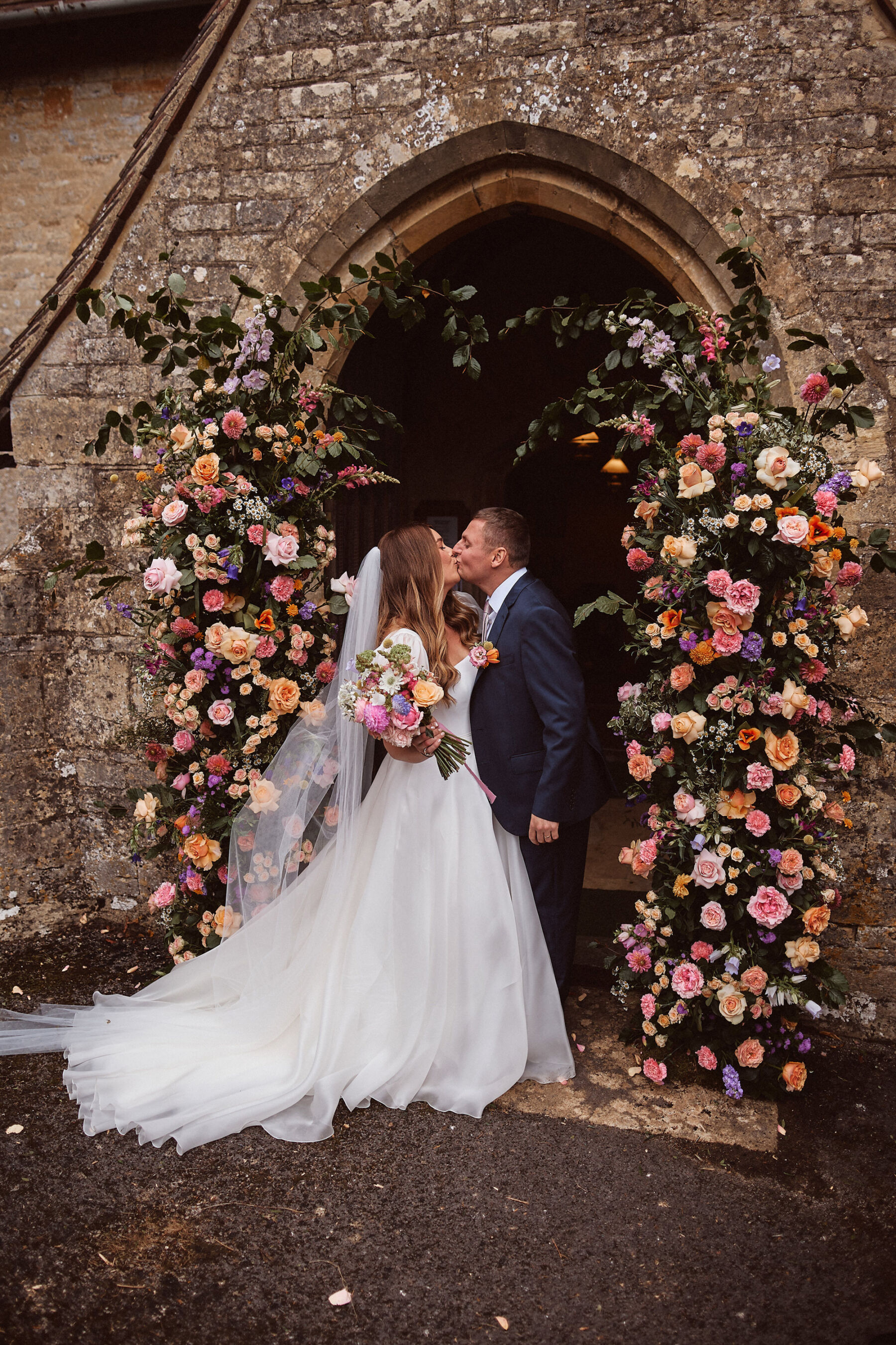 Bride and groom kissing outside a church doorway decorated with a broken floral arch.