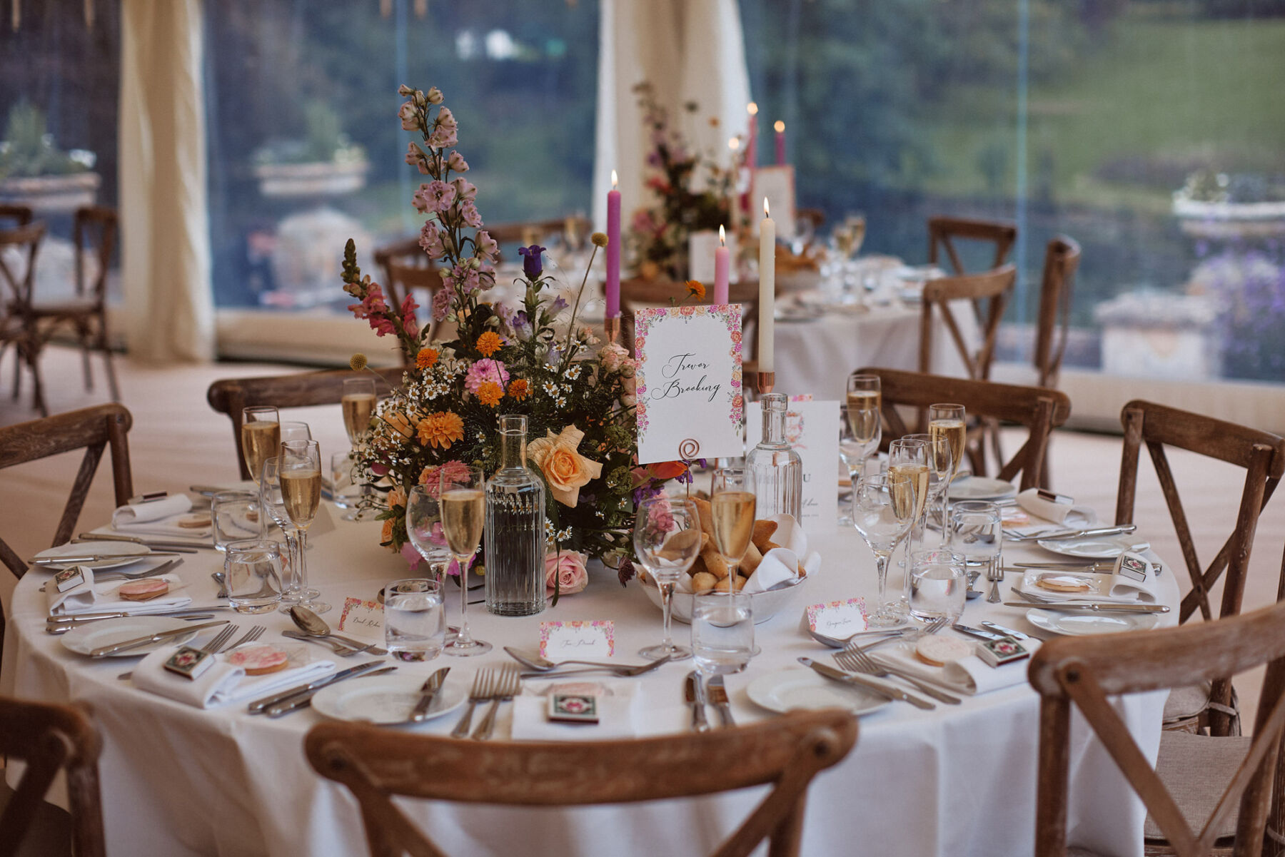 Wedding reception table with cross back wooden chairs, pink taper candles and a floral table centrepiece of bright & colourful, late summer flowers.