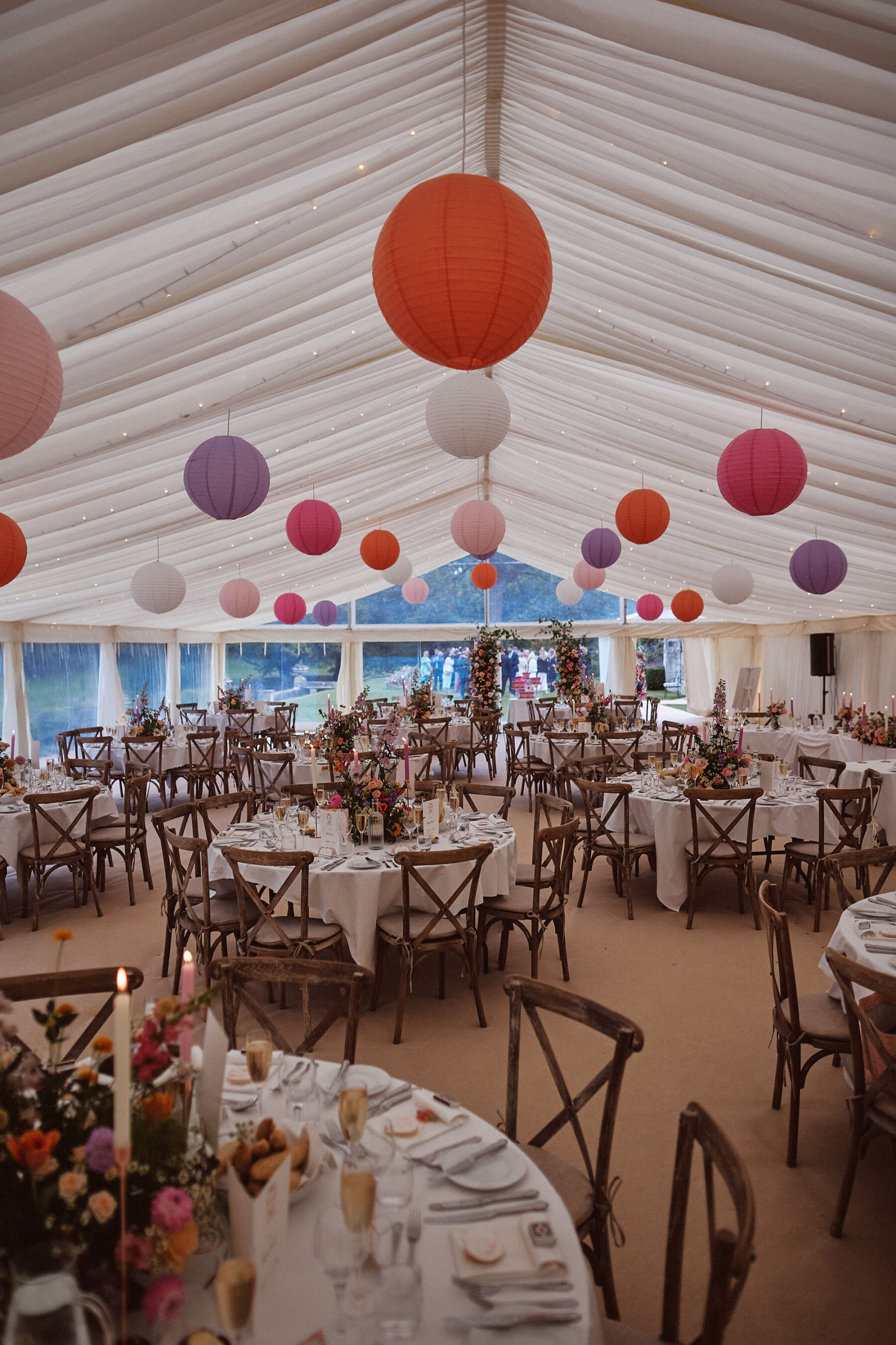 Colourful lanterns hanging within a wedding marquee. Round tables with cross back chairs and colourful floral table centrepieces.