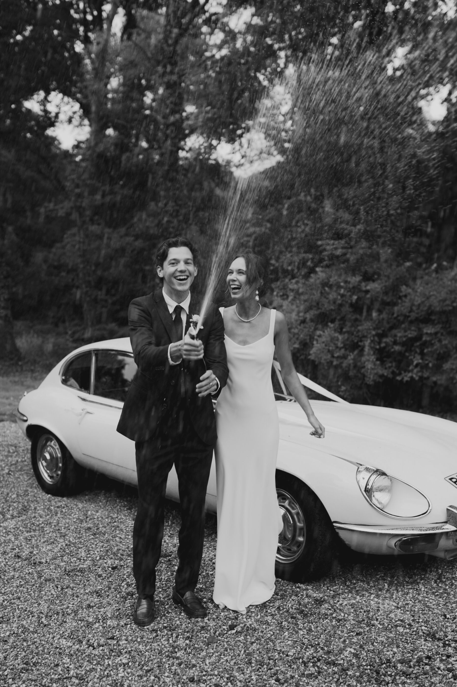 Black and white photography of a bride and groom standing by a vintage car popping a bottle of champagne.
