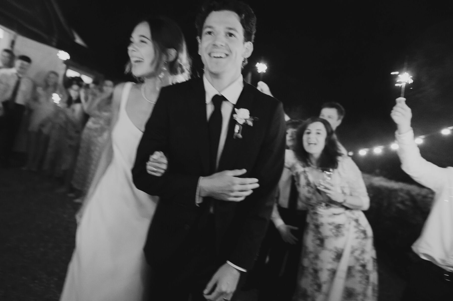 Black and white photography of bride and groom arm in arm surrounded by guests with wedding sparklers.