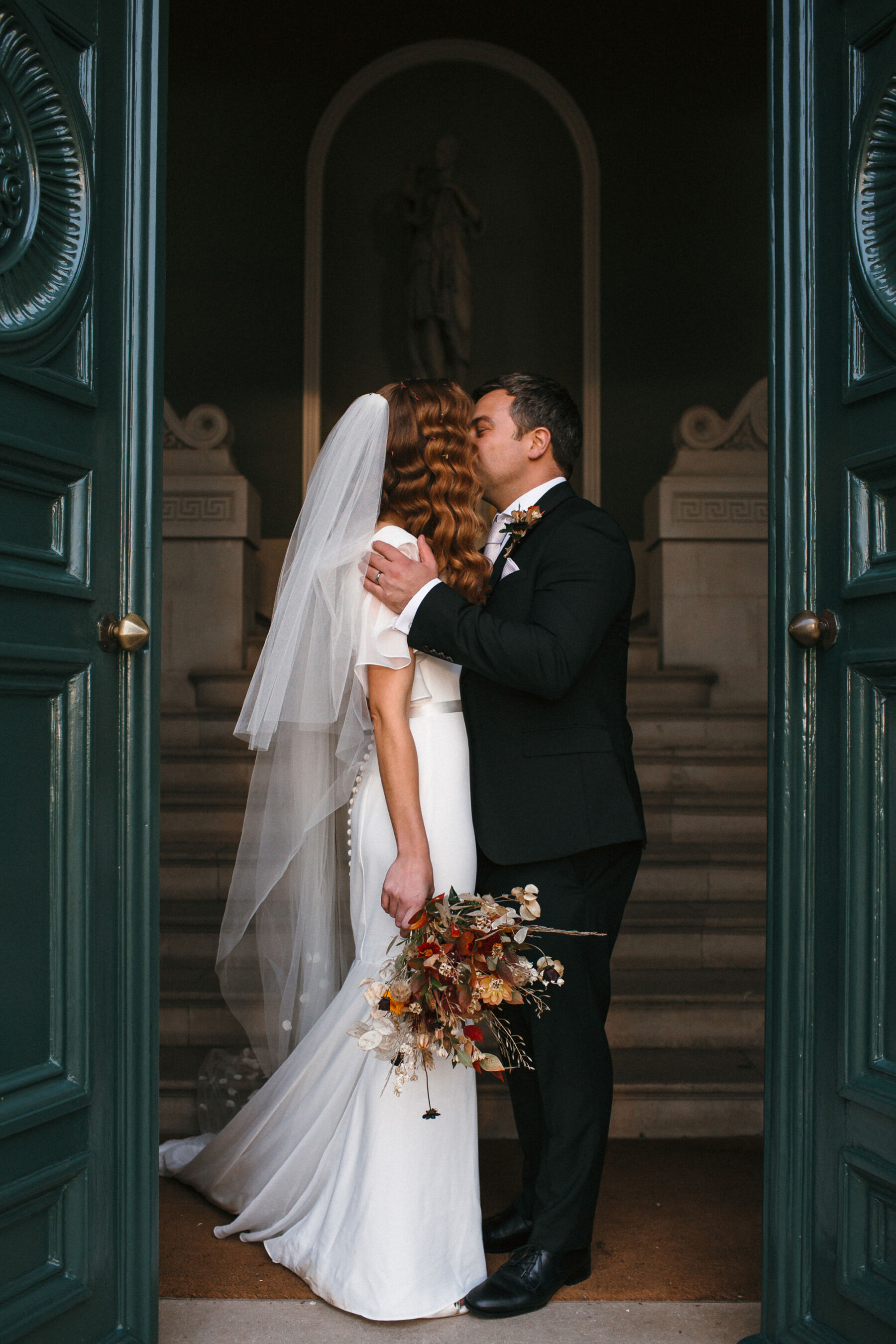 Bride with red wavy hair (marcel waves) wearing a Suzanne Neville dress, carrying an Autumn wedding bouquet & standing in the doorway of Pynes House in Devon being held and kissed by her groom.