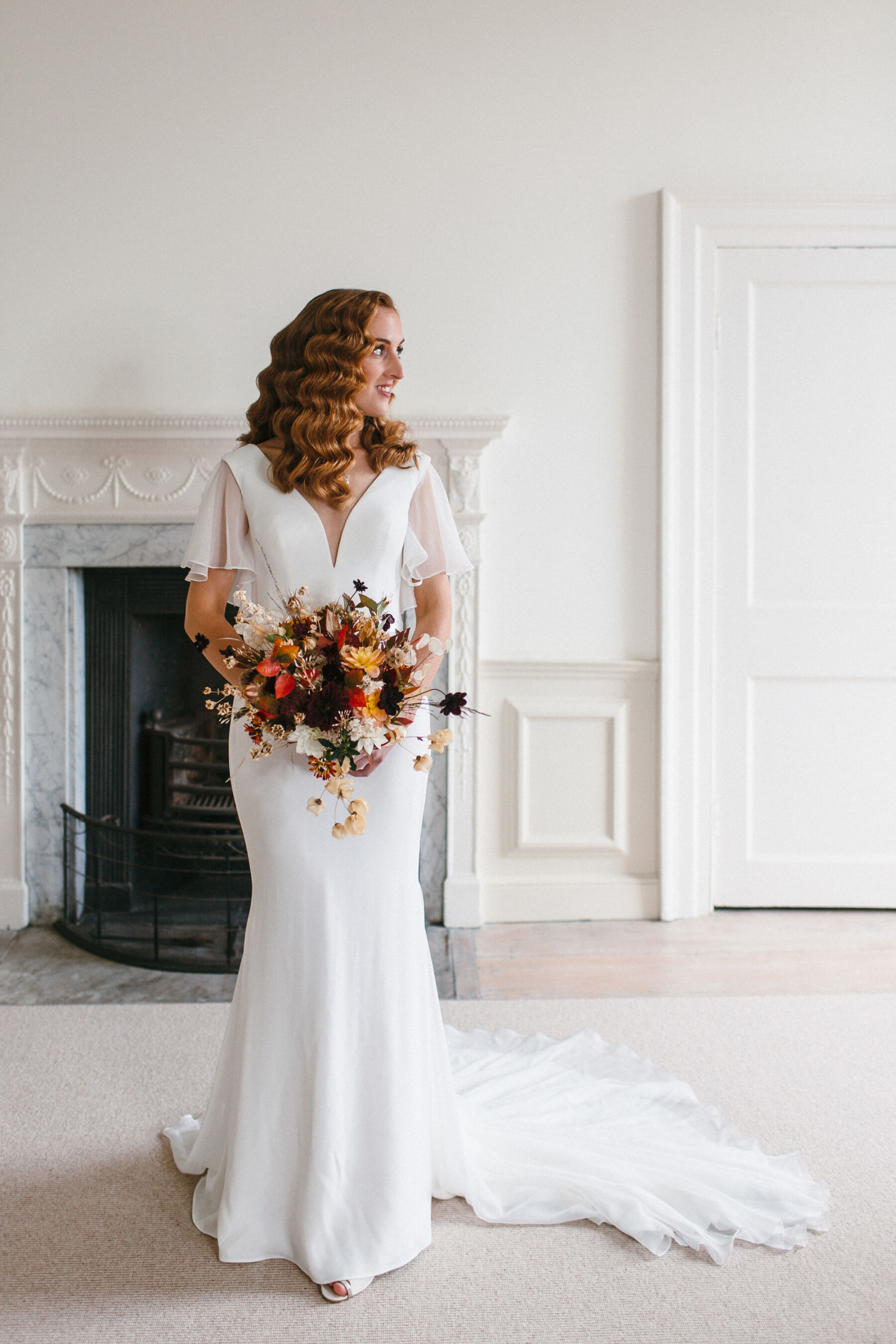 Bride wearing Suzanne Neville wedding dress with tulle sleeves. She has marcel waves in her hair and is looking to the side whilst carrying an Autumn wedding bouquet.