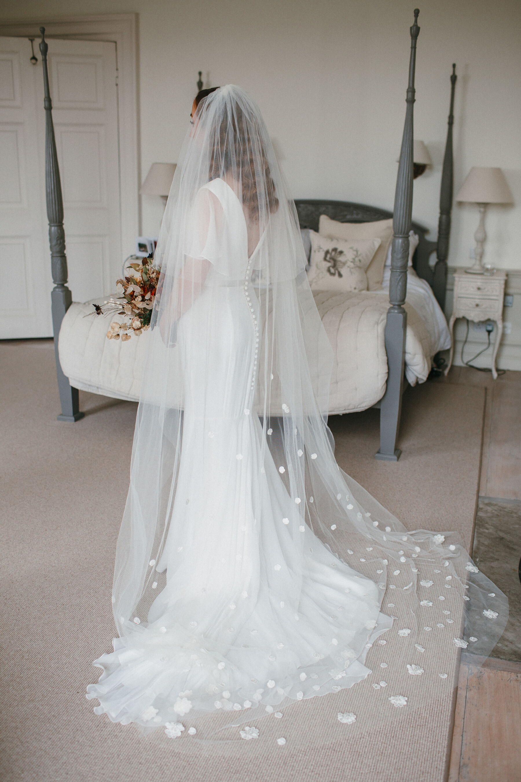 Photograph of a bride facing away from the camera wearing a long wedding veil by Suzanne Neville that is adorned with silk wedding flowers. 