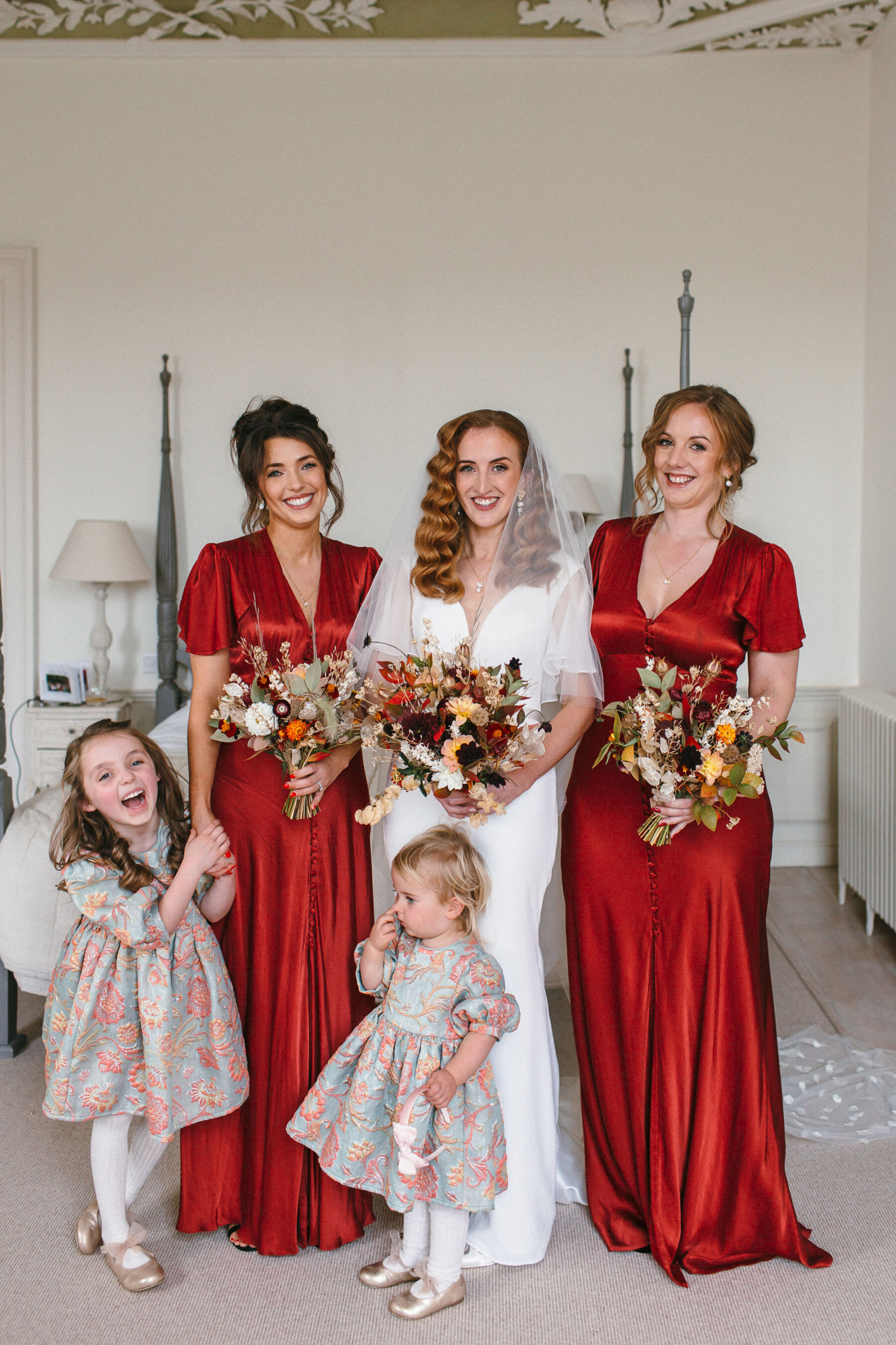 Happy smiling bride & bridesmaids. Bride wears Suzanne Neville dress and has long wavy auburn hair. Bridesmaids wear burned orange dresses by Ghost Fashion. The flower girls wear gold slip on shows with tulle bows and dresses from Next. Pynes House wedding.