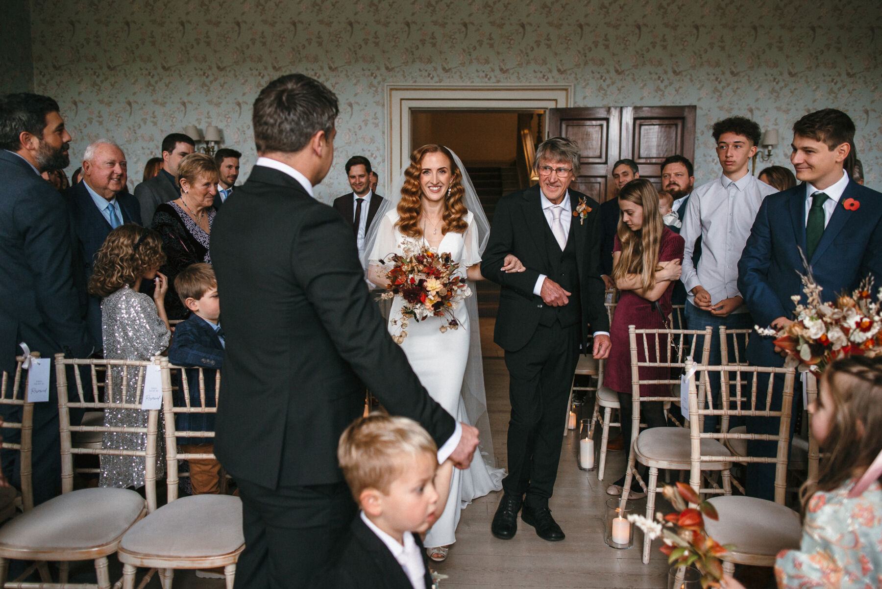 Bride with her father entering the ceremony room at Pynes House in Devon. She carries an Autumn wedding bouquet and wears long wavy hair and a Suzanne Neville wedding dress.
