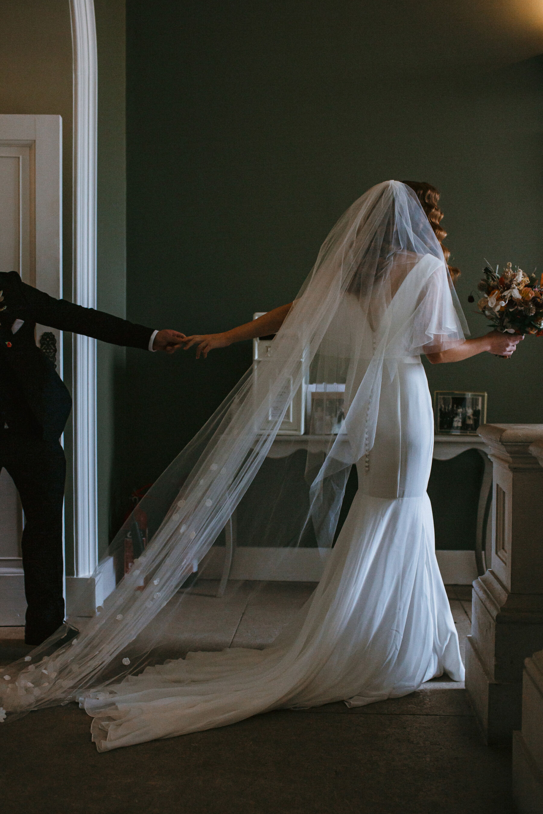 Bride in a Suzanne Neville wedding dress and long veil that trails behind her with the train of her wedding dress. Pynes House wedding, Devon.