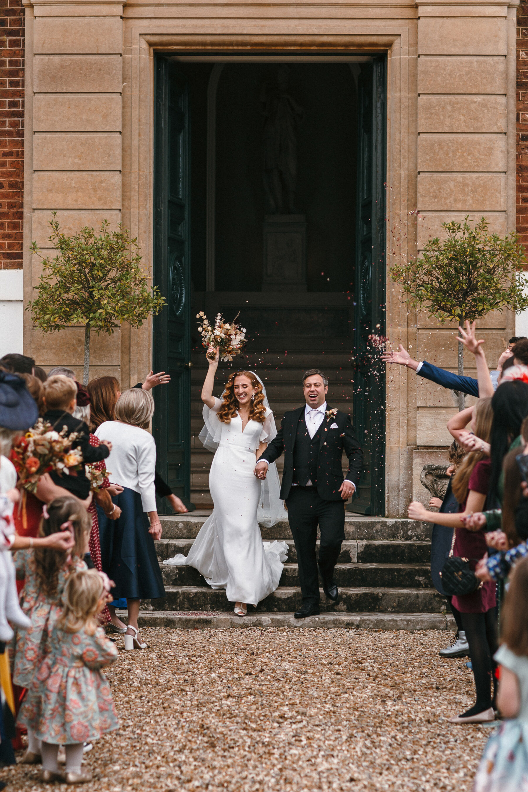 Bride and groom exiting their wedding ceremony to a shower of confetti at Pynes House in Devon. The bride wears Suzanne Neville and has long red wavy hair - she holds her bouquet up in the sky in celebration. Her groom wears a dark three piece suit.