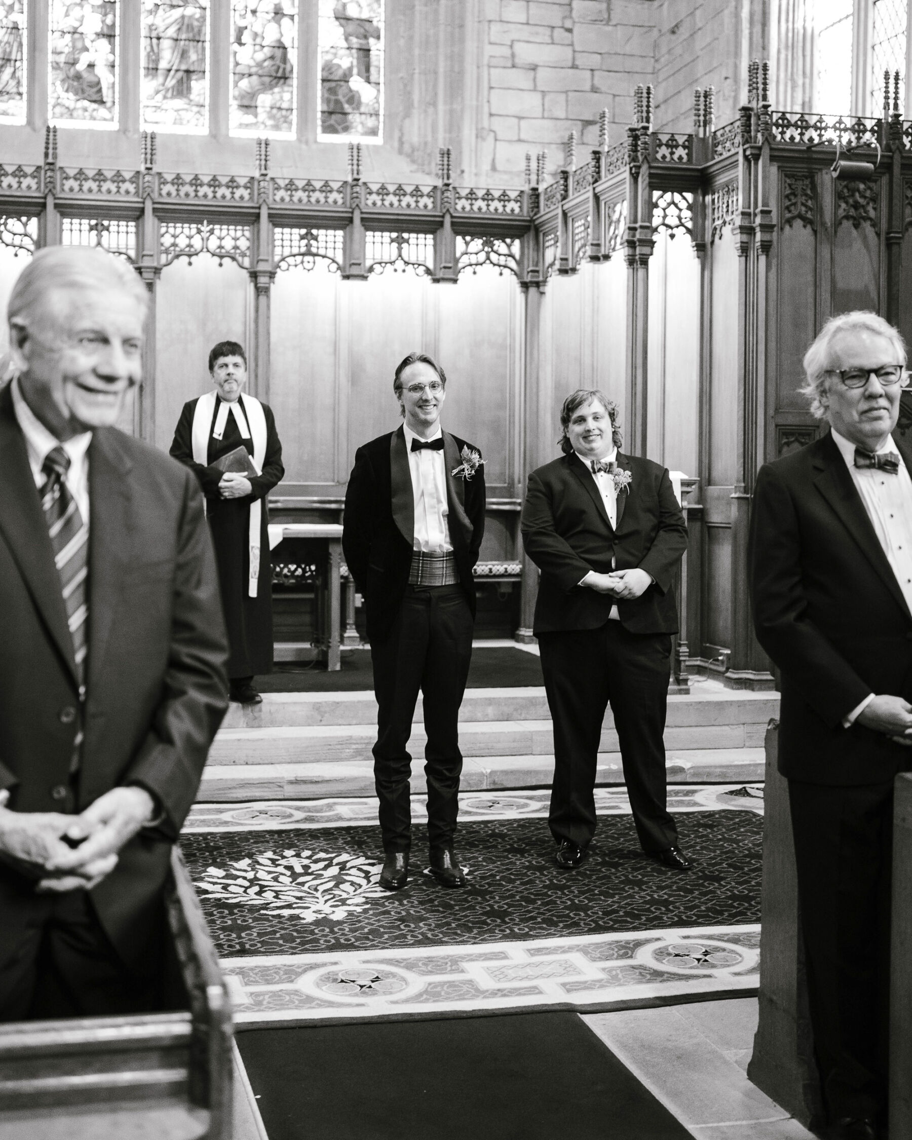 Groom in black standing inside a church smiles as he sees his bride walking down the aisle.