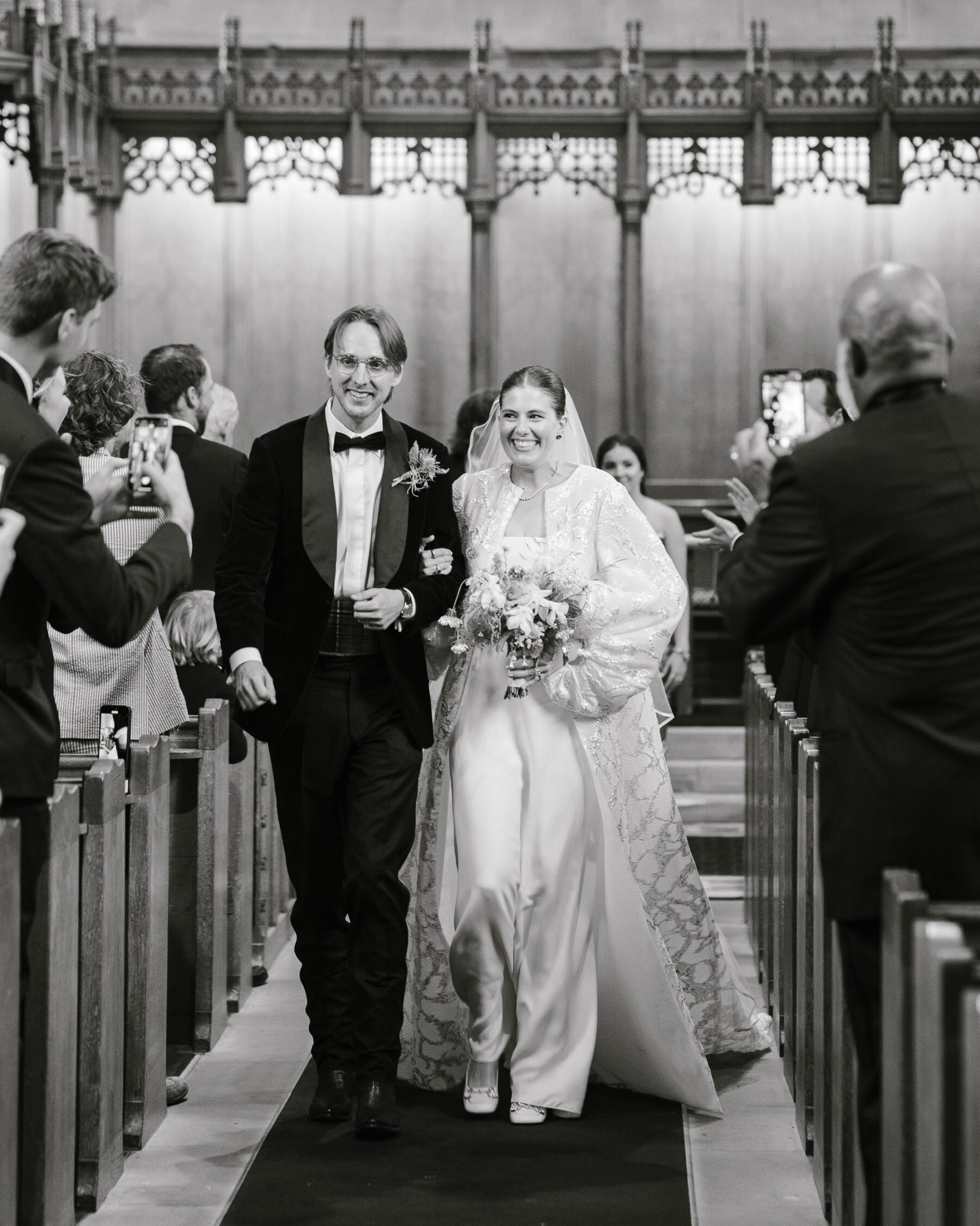 Bride in The Row dress & Jesus Peiro coat, and groom in black tie. The couple are arm in arm and smiling as they walk back down the aisle of their church wedding ceremony.