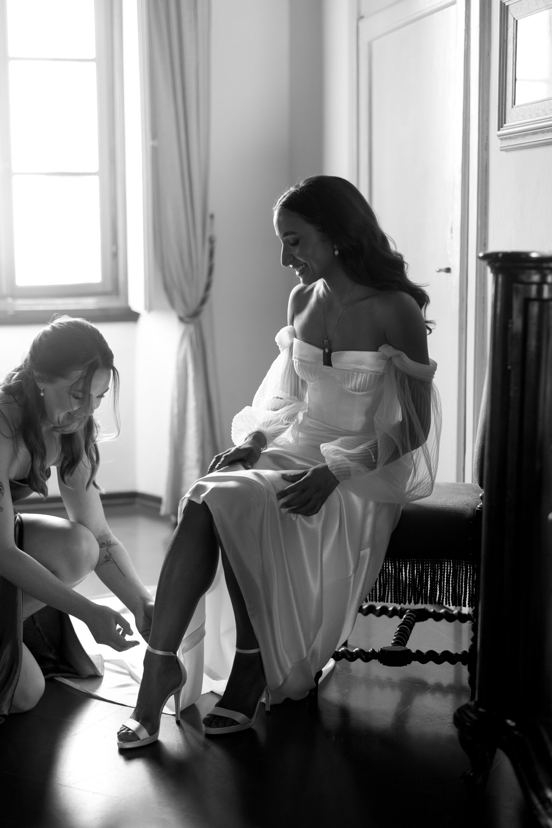 Modern bride wearing an Alena Leena wedding dress, seated whilst her bridesmaid helps her into her high heel strappy sandal wedding shoes.