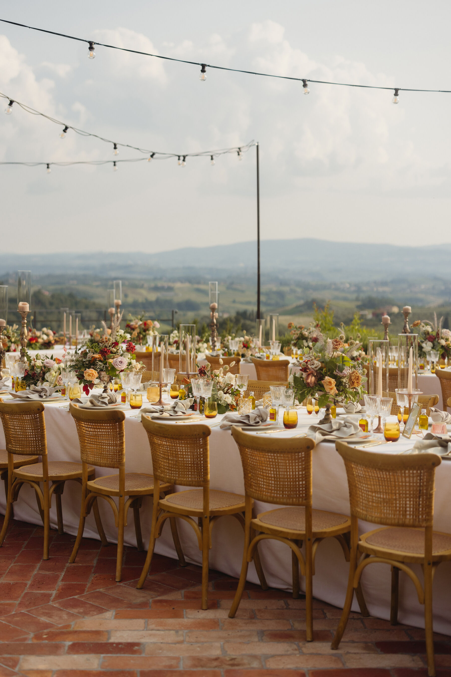 Outdoor wedding reception table setup - wedding in Italy. Wooden chairs surrounding a long table that has colourful summer floral decor in the centre and crisp white table cloths. The table has festoon lights draped above it to add ambience in the evening.
