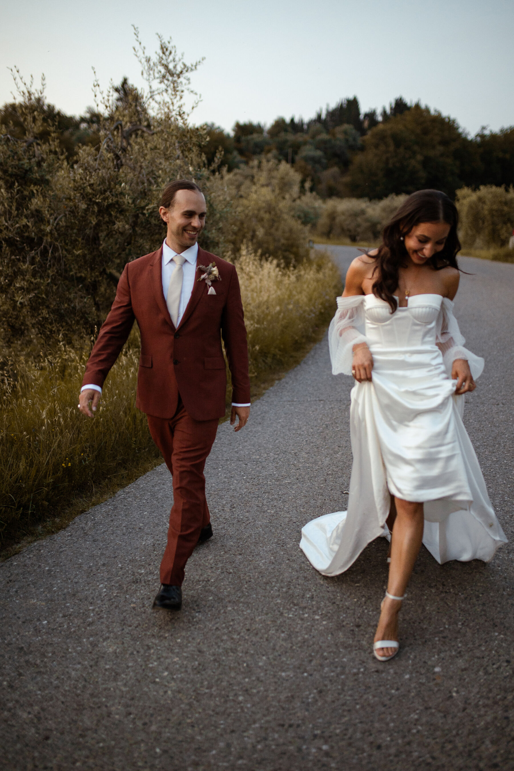 Modern bride and groom playfully walking along a stone road in Tuscay, Italy on their wedding day. She wears a modern off the shoulder Alena Leena wedding dress with detachable tulle sleeves and is raising it to bear her lower legs and high heel strappy sandal wedding shoes. Groom wears a burned orange suit by Paul Smith. 