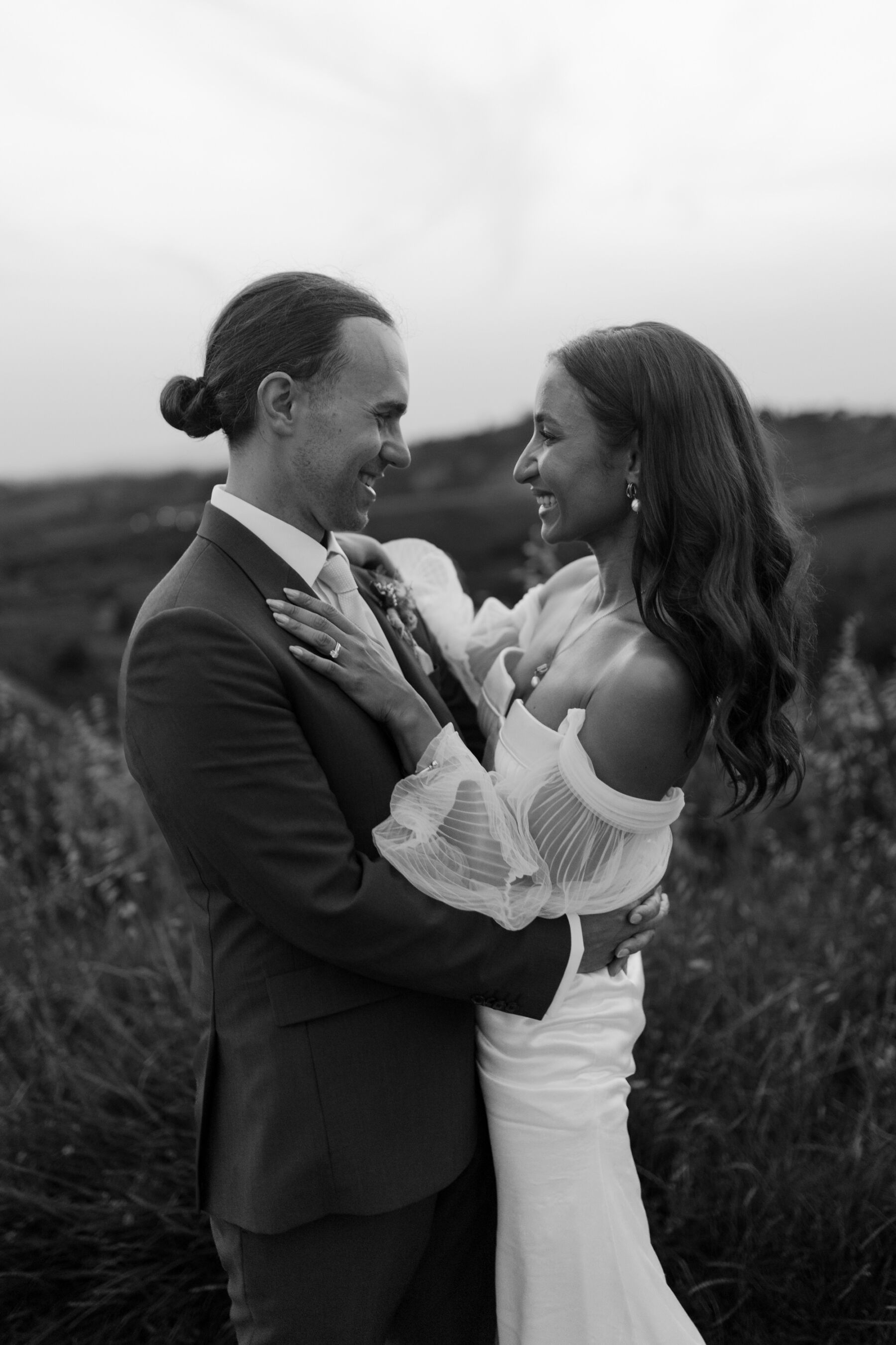 Black and white photography of a modern bride and groom intimately embracing and staring lovingly into each other's eyes. She wears a modern off the shoulder Alena Leena wedding dress with detachable tulle sleeves and is raising it to bear her lower legs and high heel strappy sandal wedding shoes. Groom wears a suit by Paul Smith. 