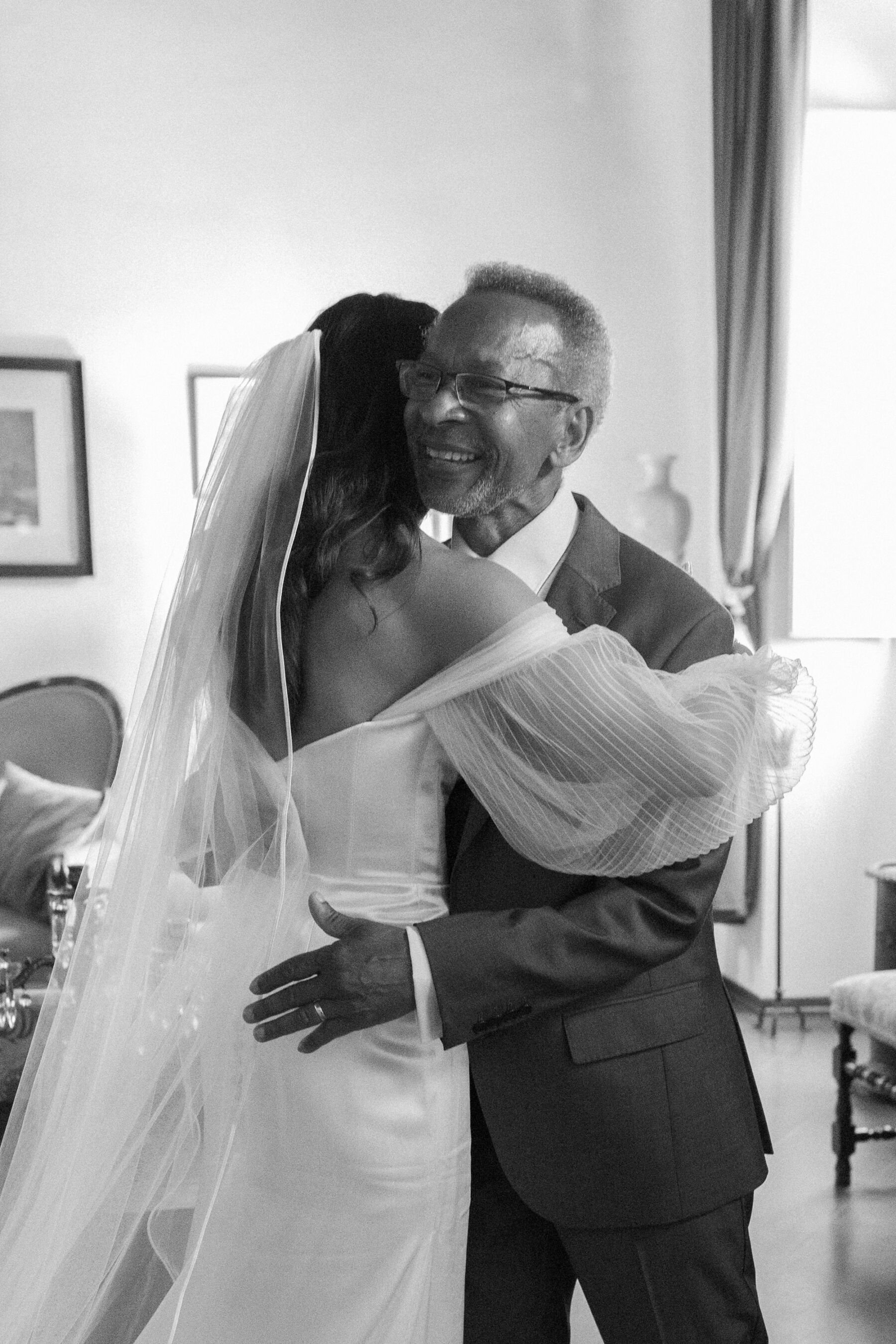 Photography of the back of a bride in an Alena Leena wedding dress, hugging her father who looks happy and joyous.