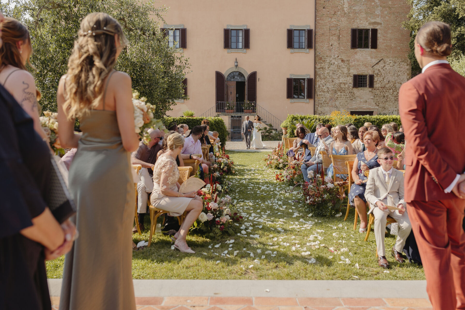 Long shot of a bride arm in arm with her father arriving at her outdoor Italian wedding ceremony. The seated guests are turning to look at her, in her Alena Leena wedding dress.