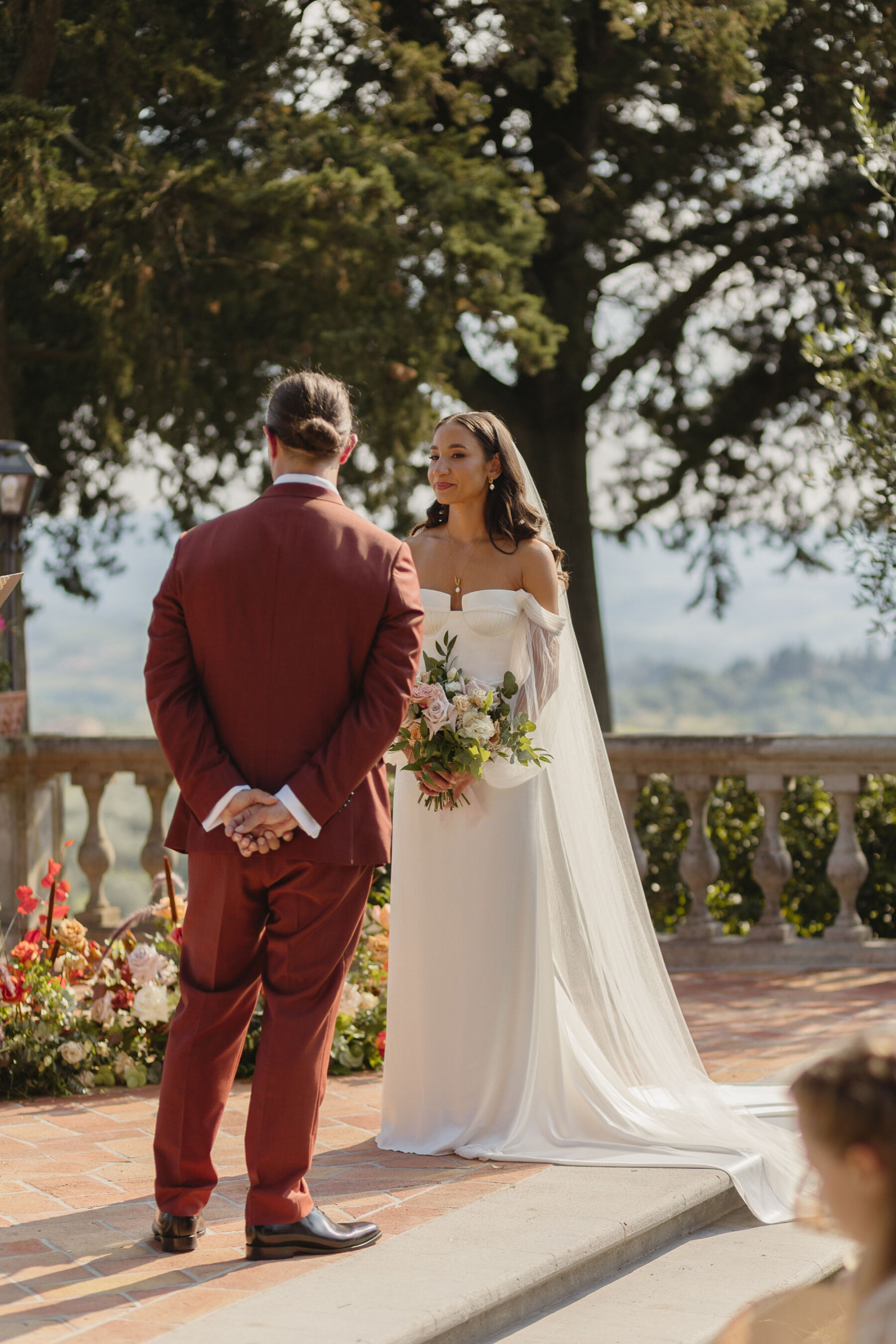 Bride and groom standing during their outdoor wedding ceremony in Tuscany, Italy. The bride wears an Alena Leena wedding dress & the groom wears a burnt orange Paul Smith suit. The bride carries a colourful and elegant bouquet at waist level.
