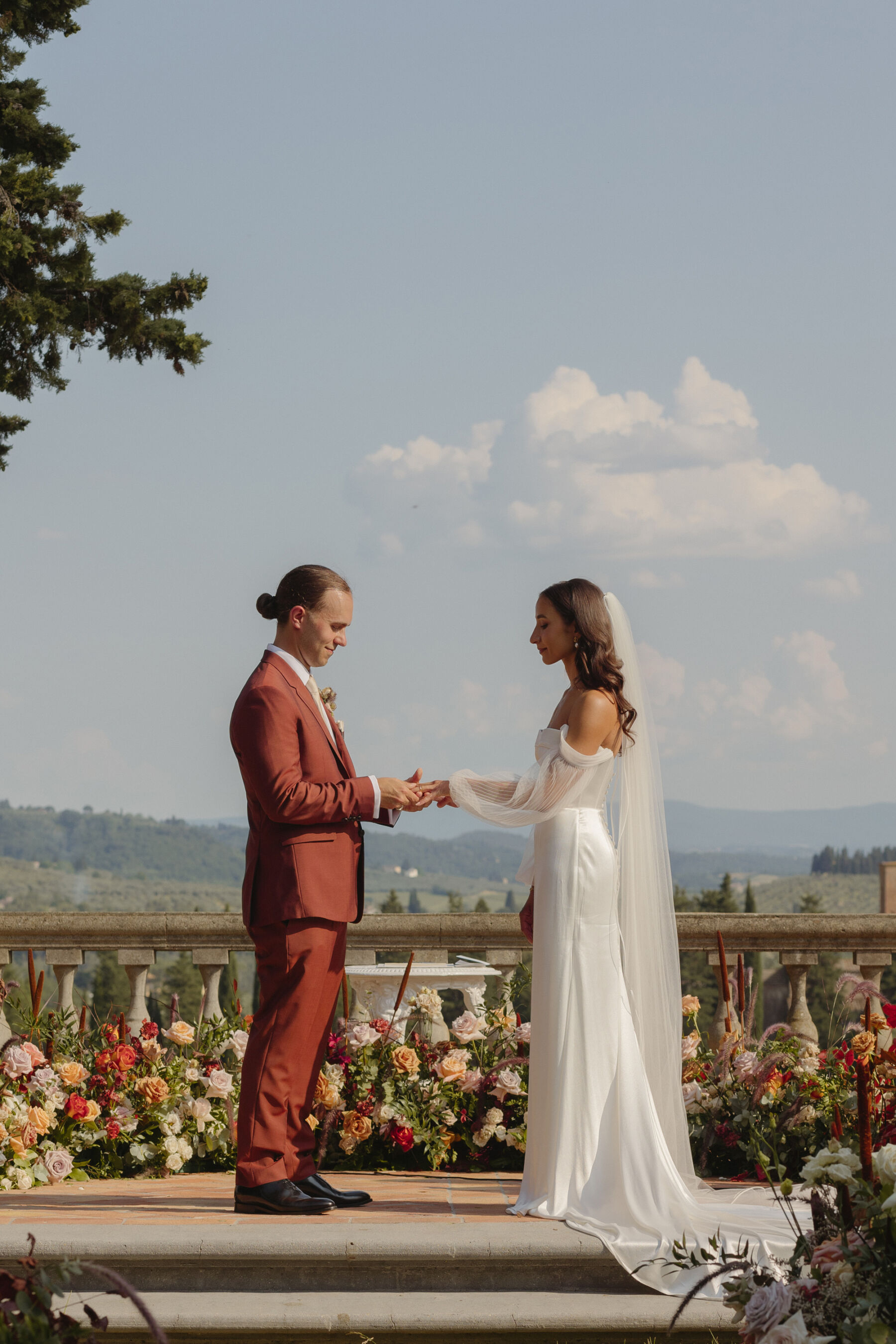 Bride and groom holding hands during their outdoor wedding ceremony in Tuscany, Italy - rolling hills visible in the background. The bride wears an Alena Leena wedding dress & the groom wears a burnt orange Paul Smith suit. The bride carries a colourful and elegant bouquet at waist level.