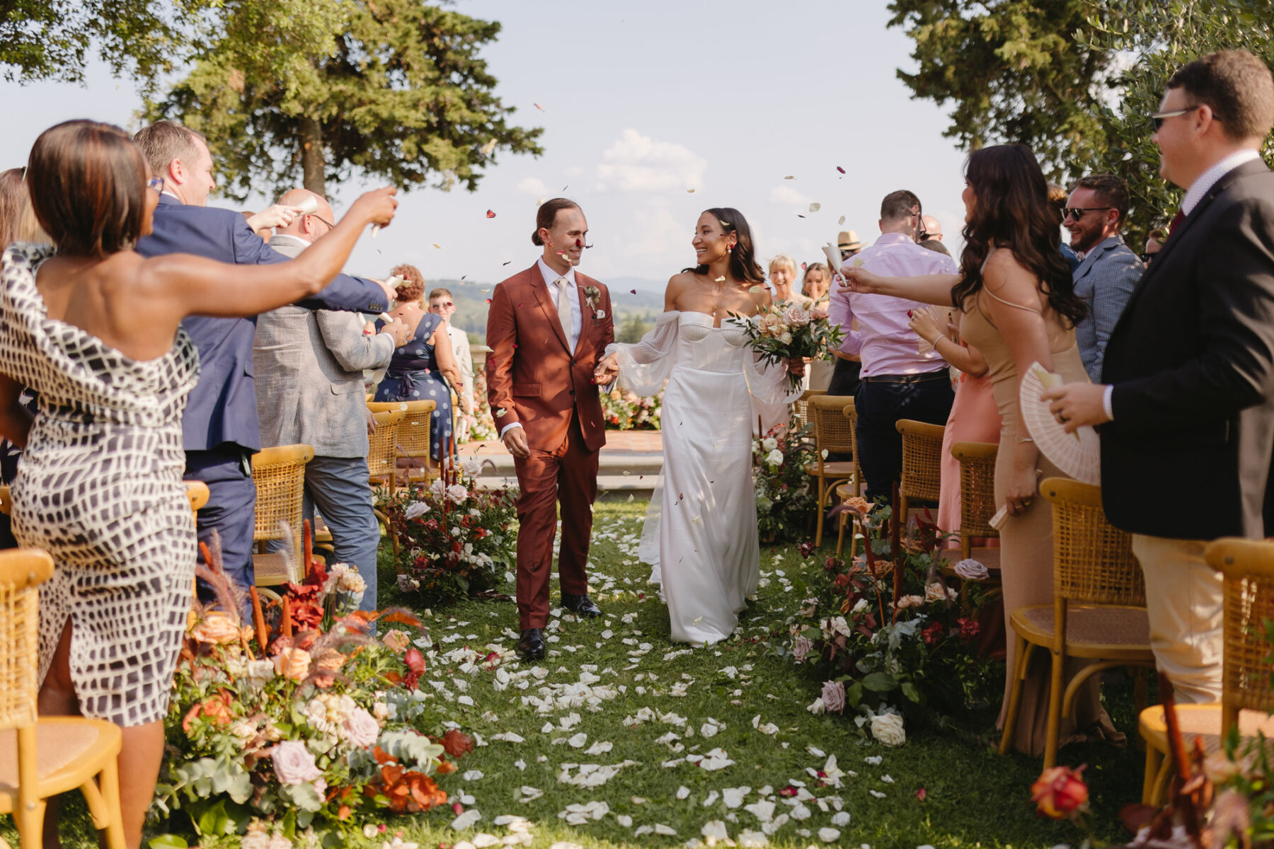 Happy and joyous bride and groom exiting down the aisle of their outdoor Italian wedding ceremony. He wears a burnt orange Paul Smith suit, the bride who is leaning back in the embrace, wears an Alena Leena wedding dress. The aisle is strewn with rose petals and the guests are showering the couple with confetti. Sunny wedding in Tuscany.