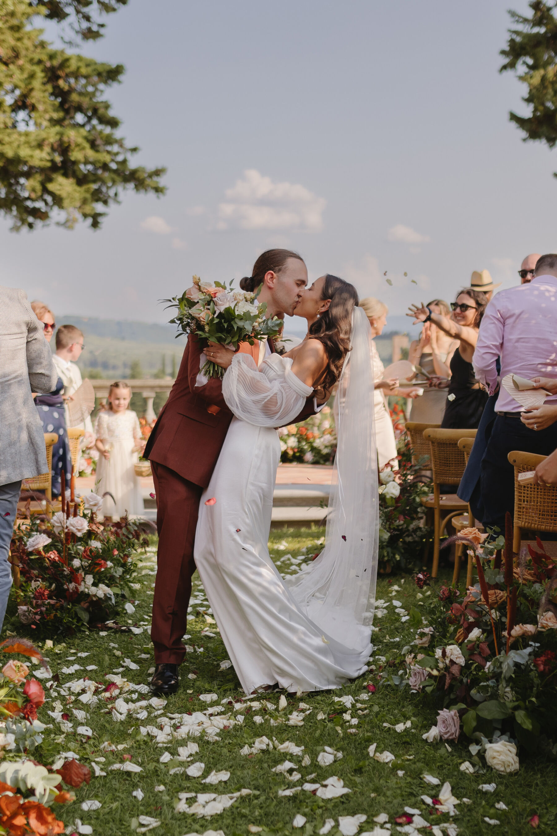 Bride and groom embracing whilst exiting down the aisle of their outdoor Italian wedding ceremony. He wears a burnt orange Paul Smith suit, the bride who is leaning back in the embrace, wears an Alena Leena wedding dress. The aisle is strewn with rose petals and the guests are showering the couple with confetti. Sunny wedding in Tuscany.
