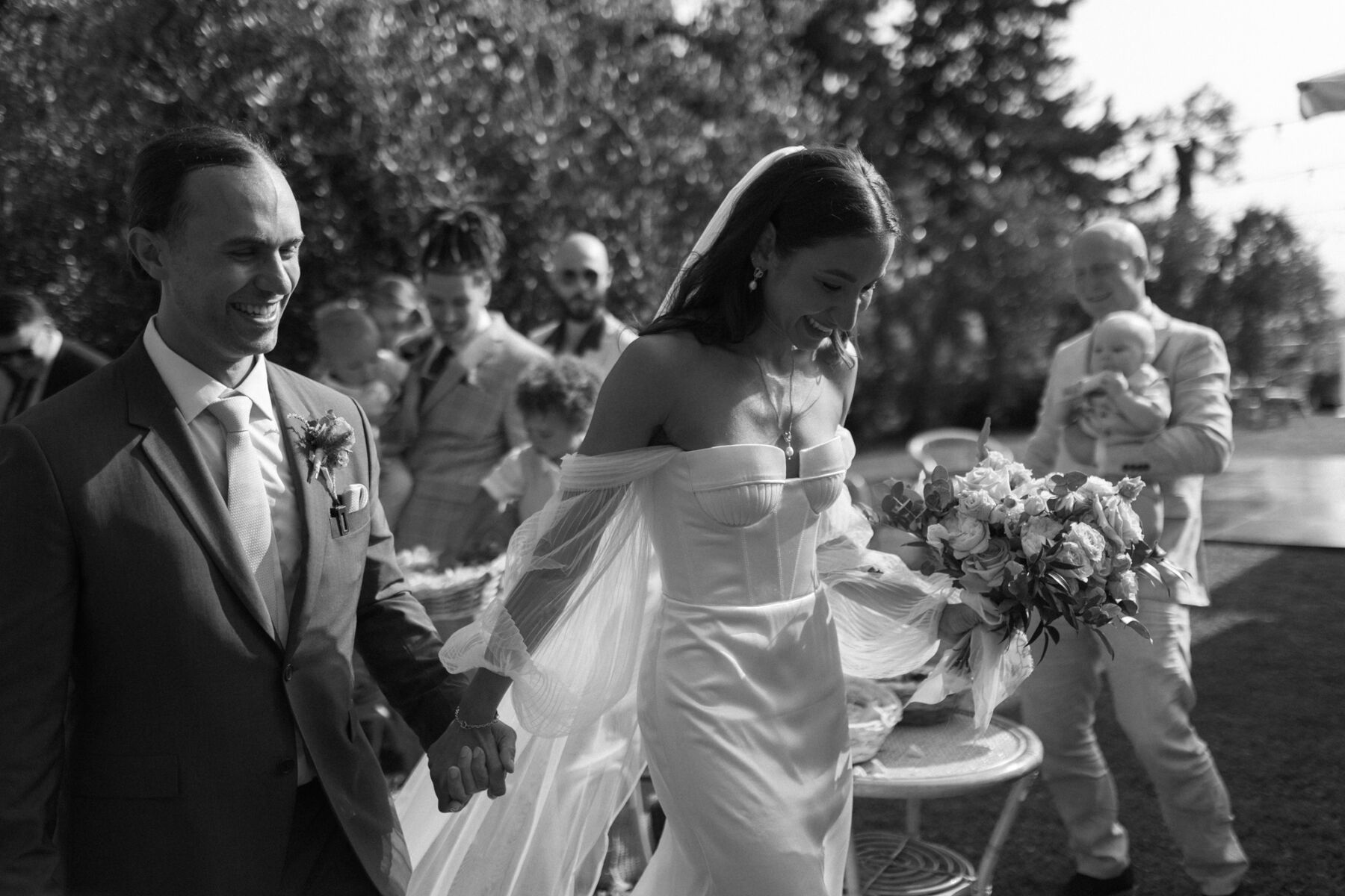 Black & white wedding photograph of bride and groom exiting their Italian wedding. She wears an off the shoulder gown by Alena Leena.