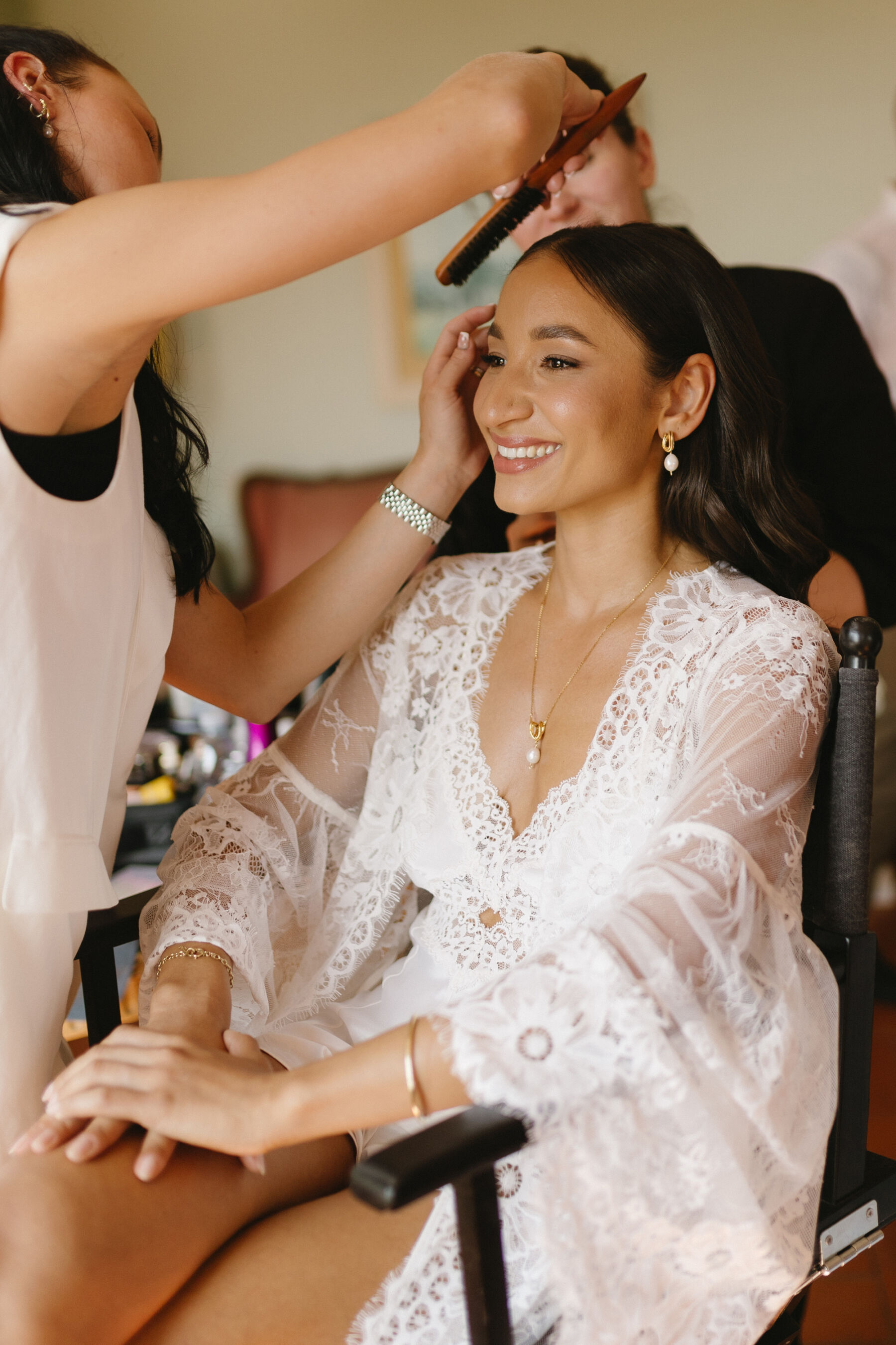 Bride in tulle and lace getting ready on the morning of her wedding, having her hair and makeup done. She wears matching modern pearl & gold earrings and necklace.