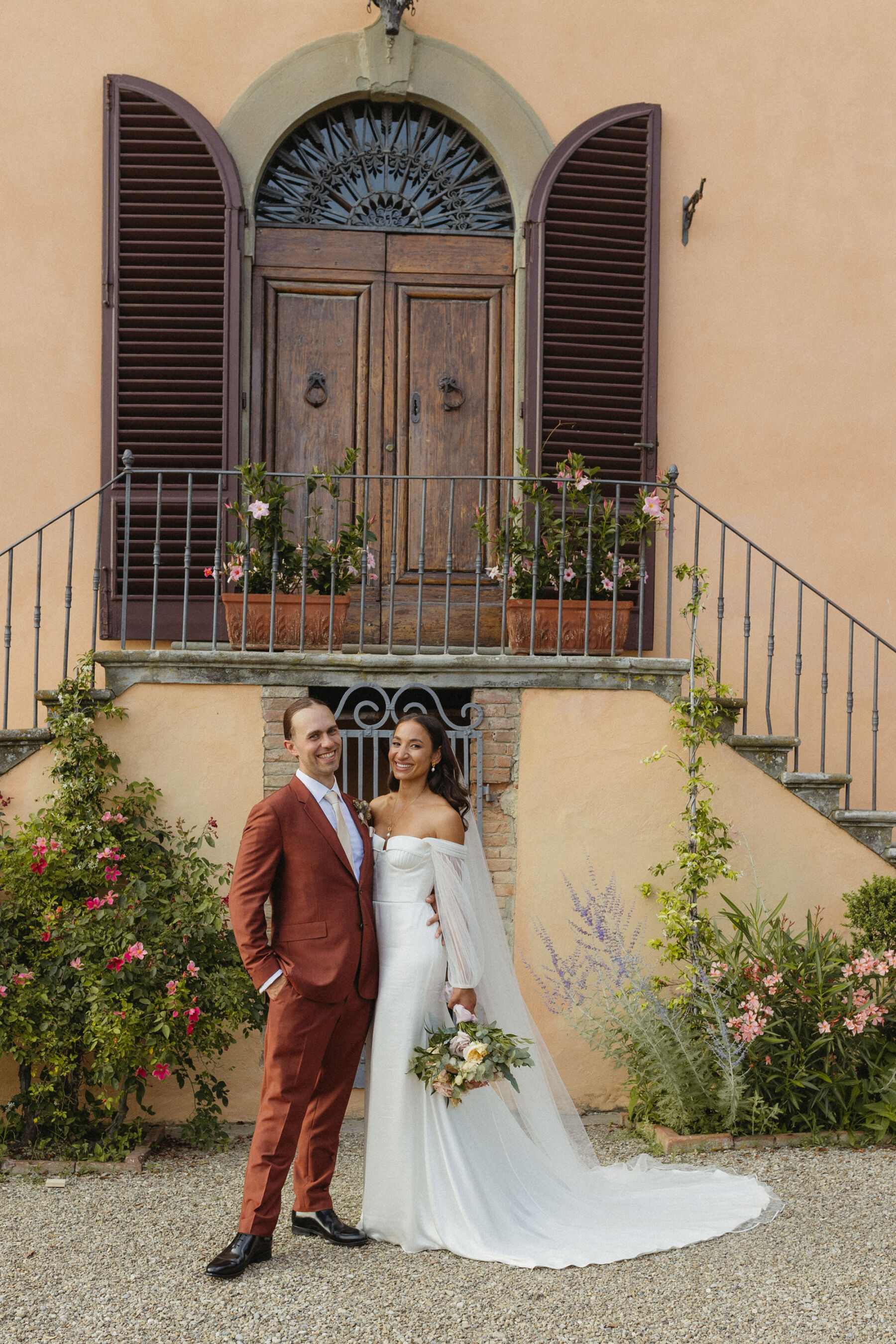 Newlywed bride and groom standing outside Villa Il Pozzo in Tuscay, Italy. He wears a burnt orange Paul Smith suit, the bride who is leaning back in the embrace, wears an Alena Leena wedding dress. 
