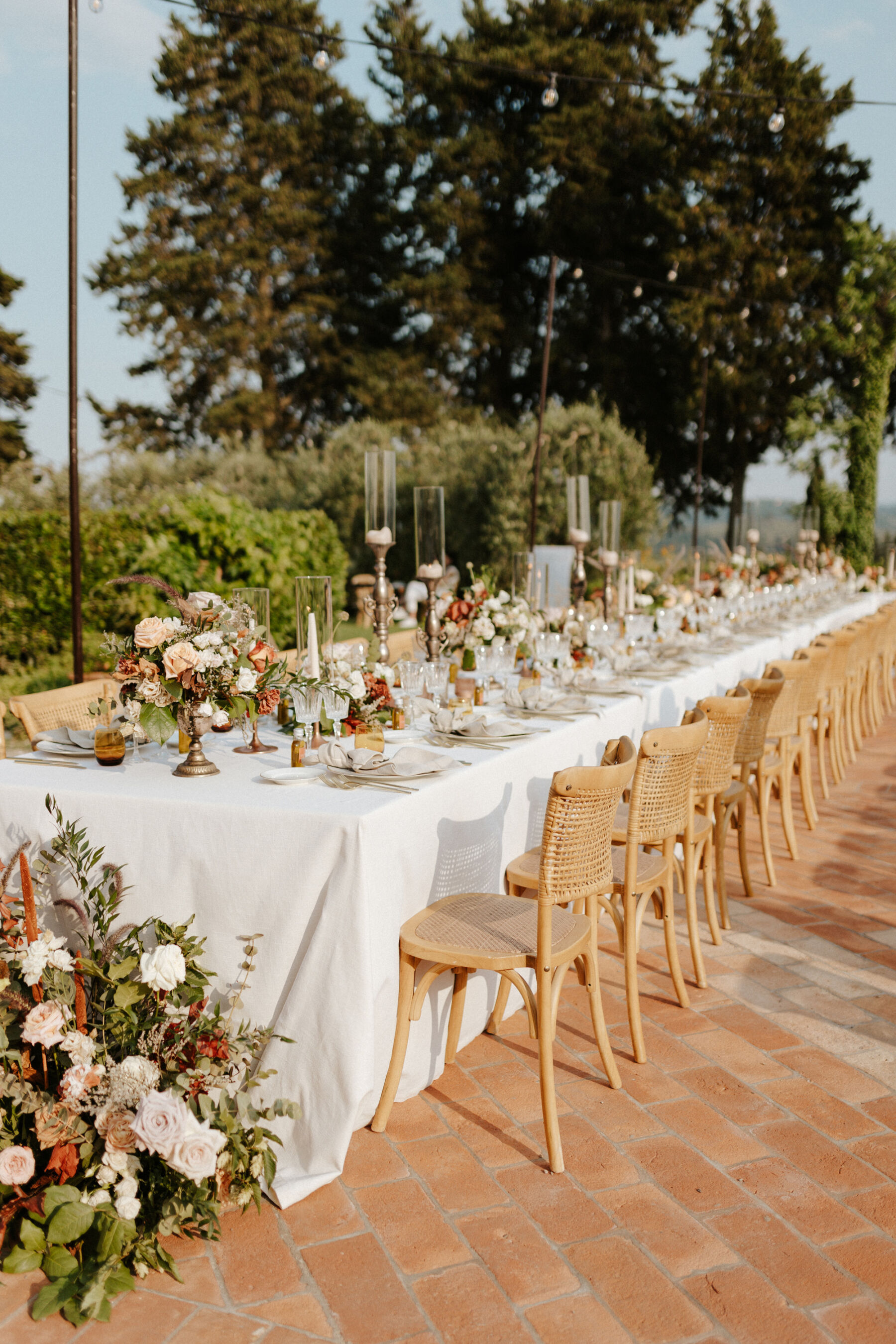 Outdoor wedding reception table setup - wedding in Italy. Wooden chairs surrounding a long table that has colourful summer floral decor in the centre and crisp white table cloths. 